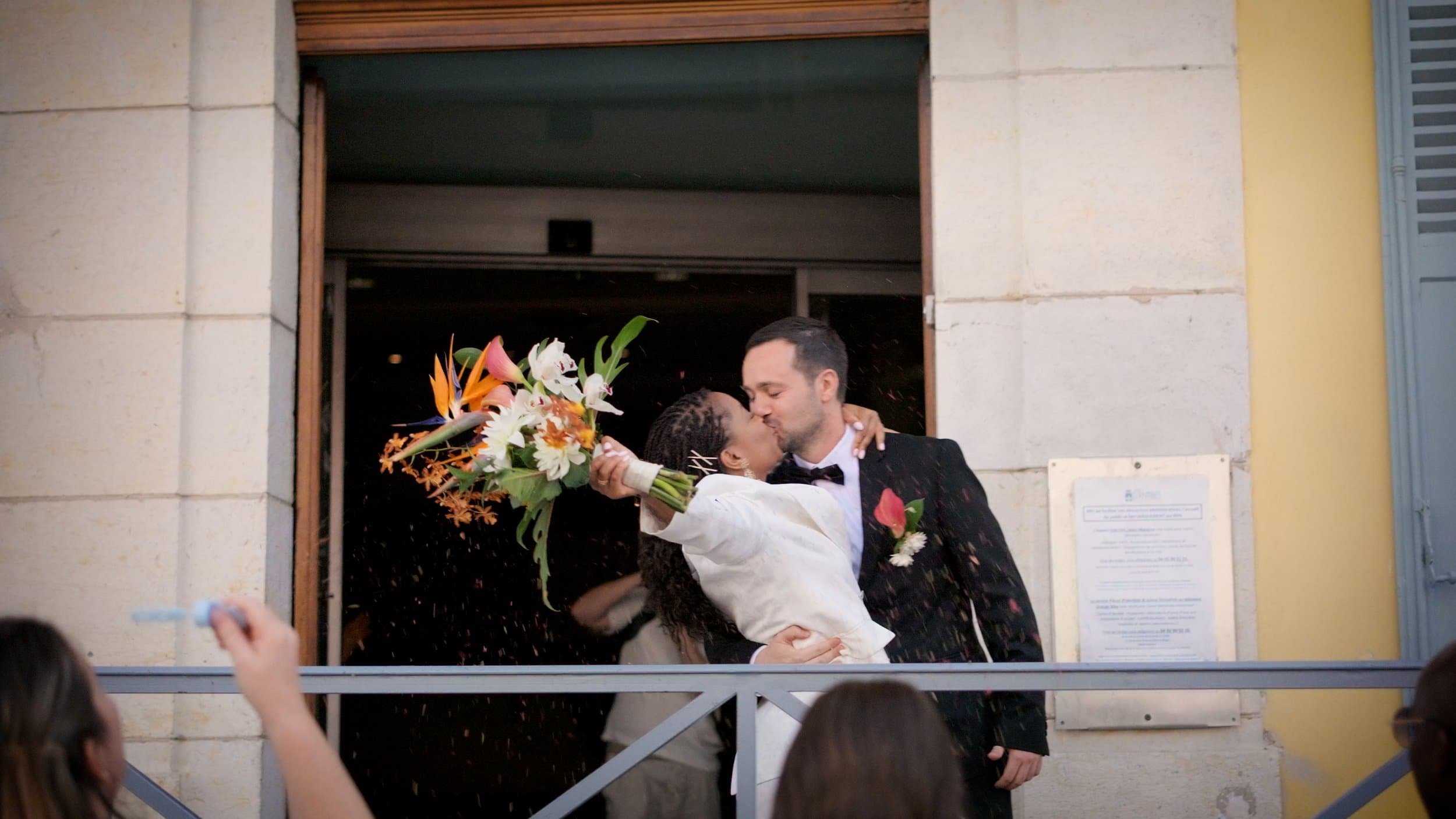 Couple de mariés s'embrassant sous une pluie de fleurs, la mariée tenant un bouquet coloré, dans une célébration de mariage en plein air.