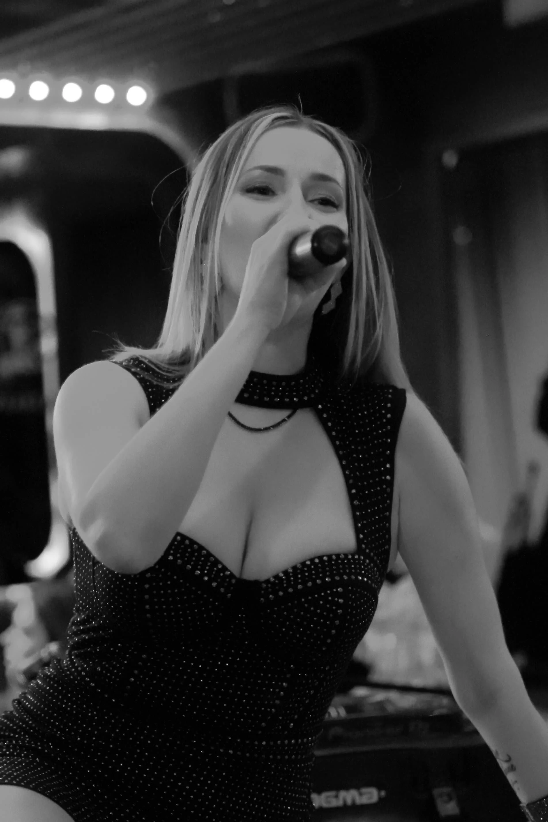 Une femme chante dans un micro, portant une robe noire à pois avec un décolleté profond, dans une ambiance de bar ou de scène.