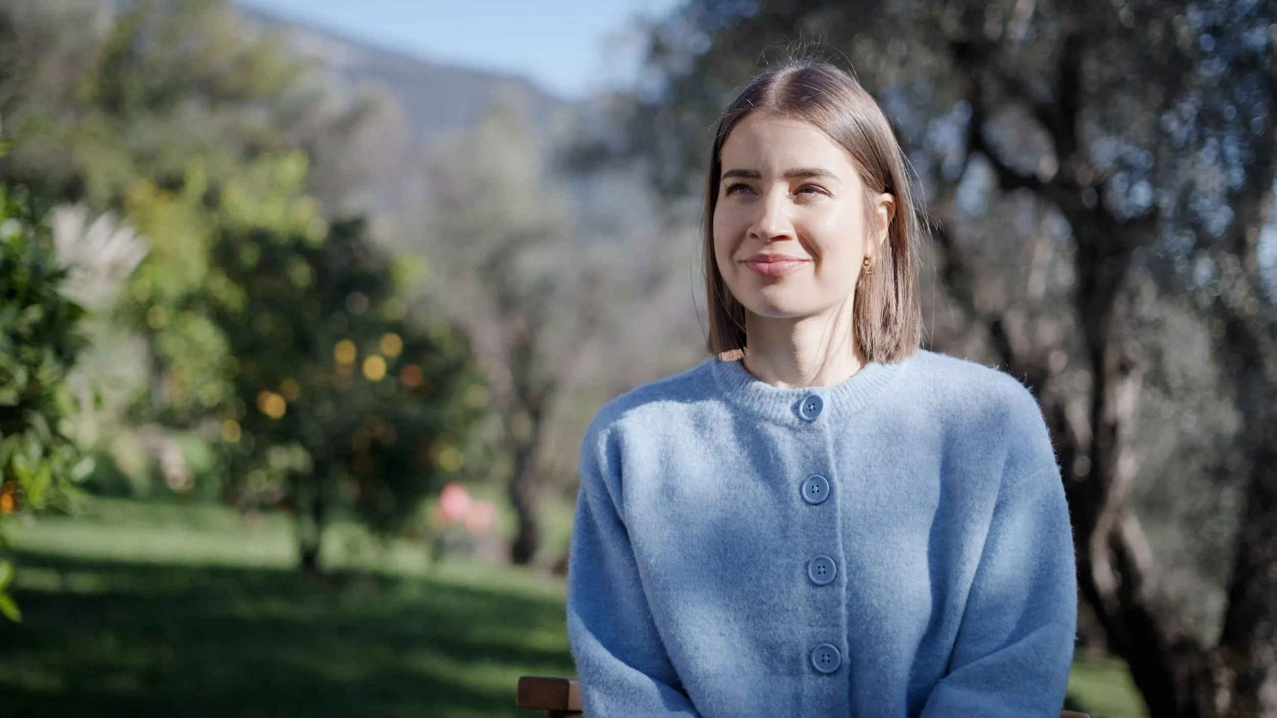 Jeune femme souriante assise dans un parc, portant un pull bleu, avec des arbres en arrière-plan.