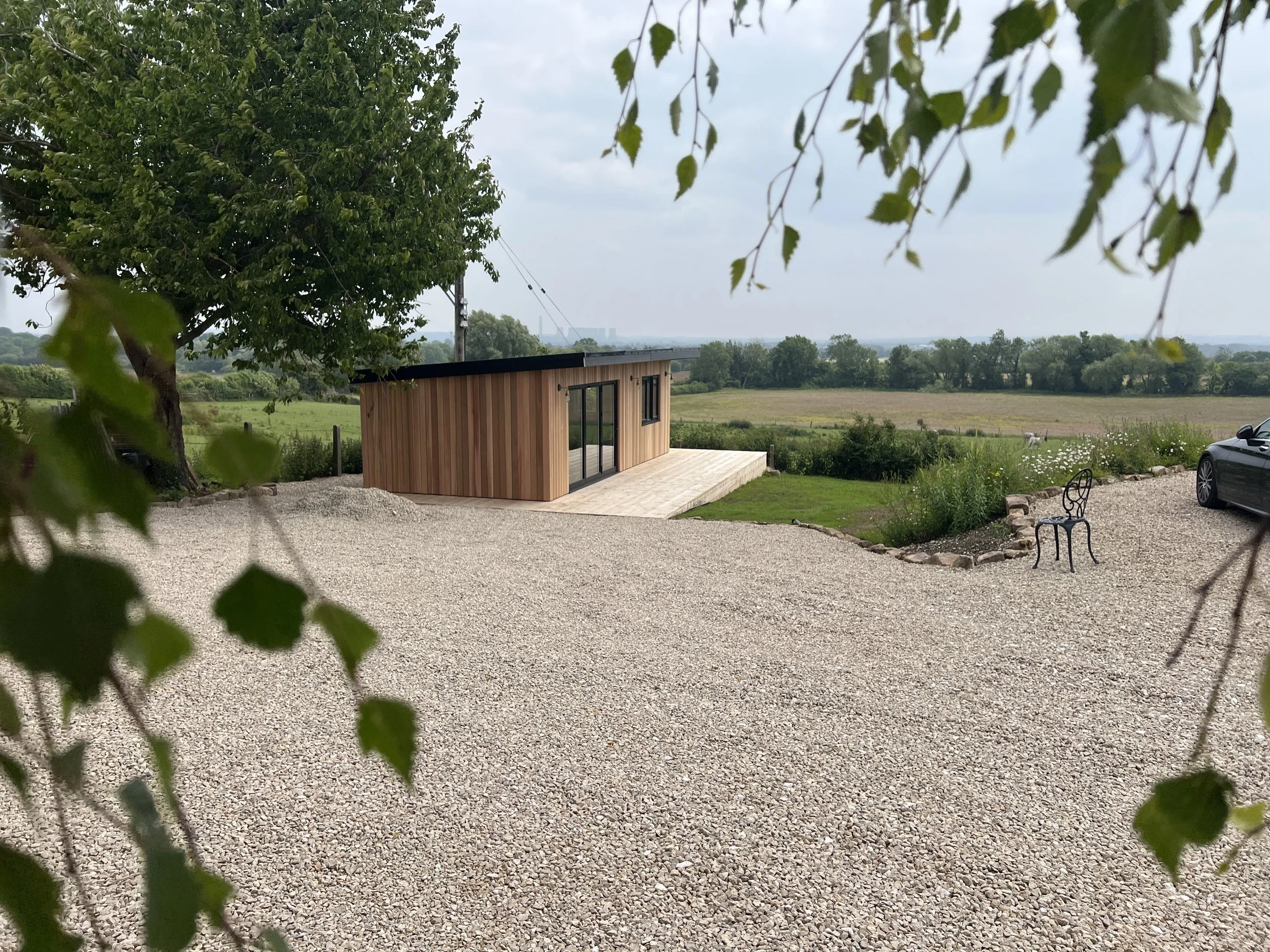 A cedar clad garden room or cabin with a black roof, located in a rural area with open fields and greenery. There is a gravel driveway with some trees and bushes around.