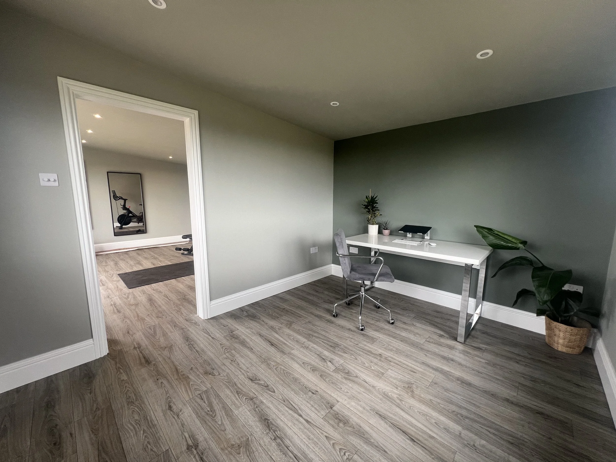 A home office with a white desk, grey chair, and potted plants against a green wall, adjacent to a room with exercise equipment.