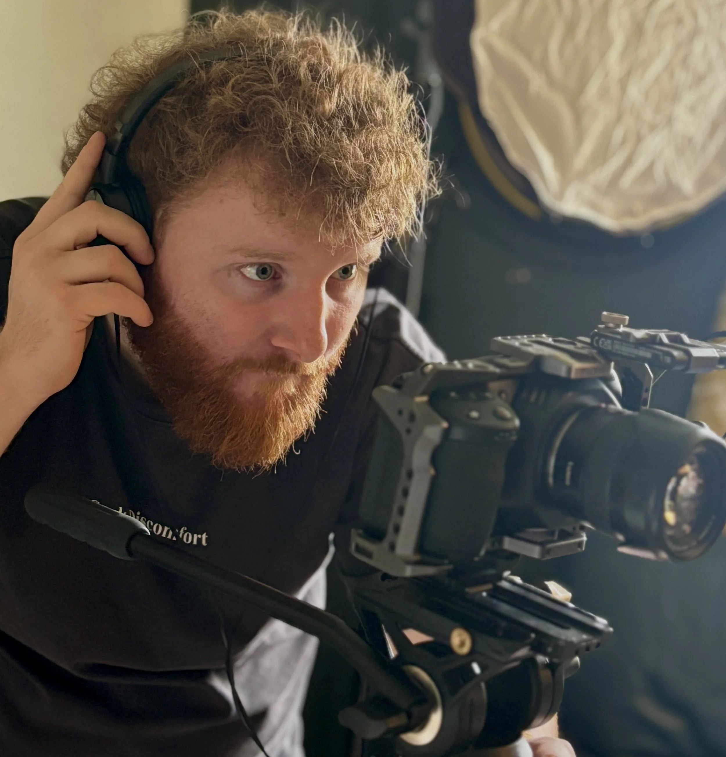A man with curly red hair and a beard is adjusting a camera mounted on a stabilizer rig, wearing headphones, in a indoor setting.