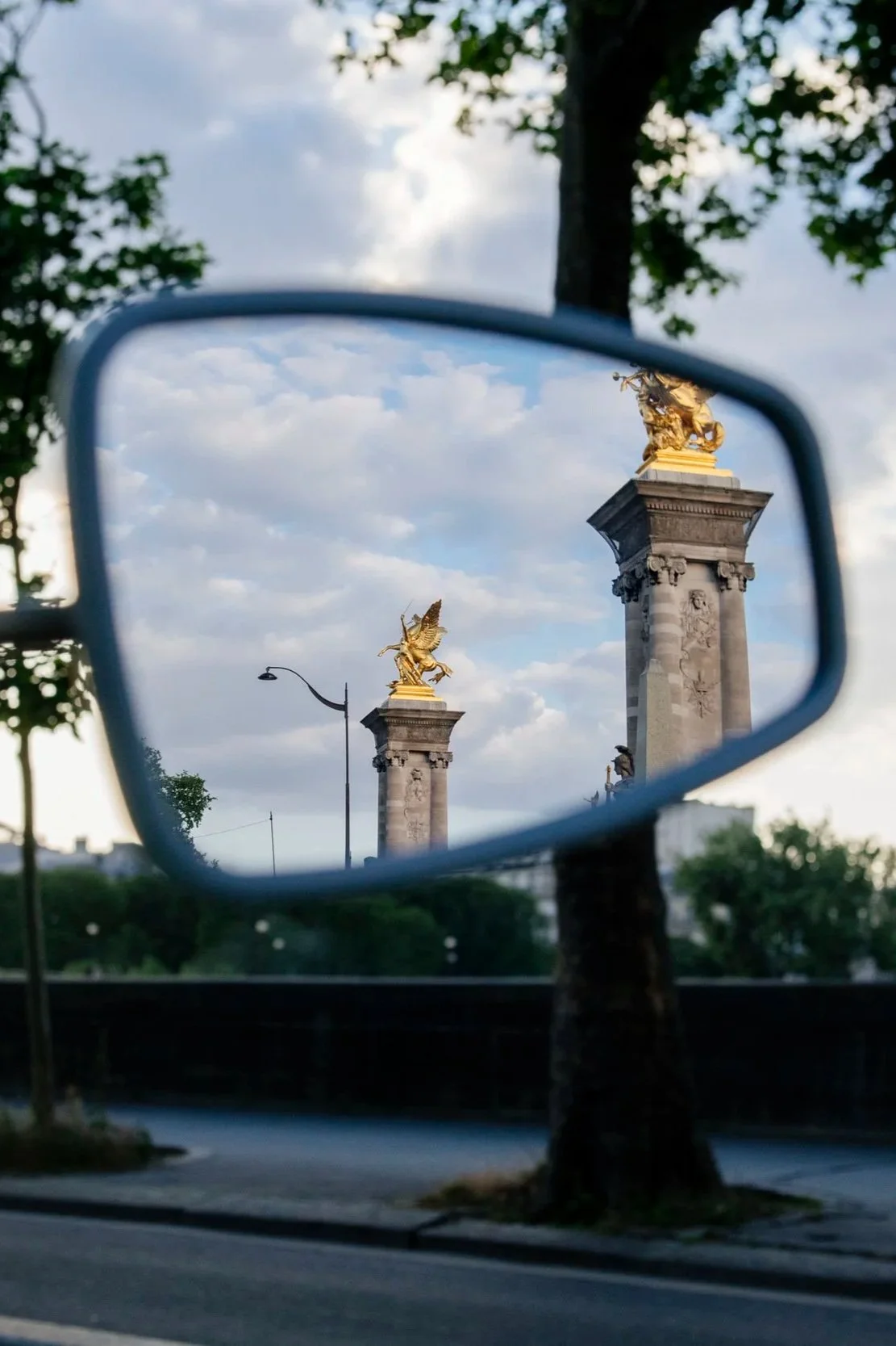 Reflet des colonnes avec des statues dorées dans un miroir de voiture, arbres, nuit et ville en arrière-plan.