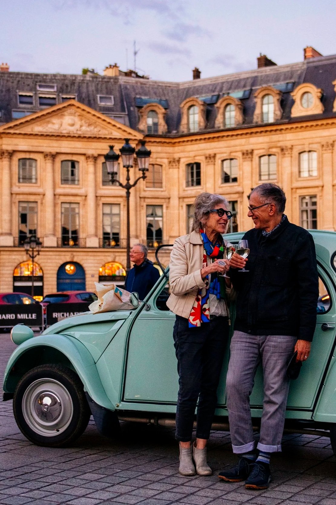 A couple standing next to a vintage green car in a European city square, holding wine glasses and smiling at each other during sunset.