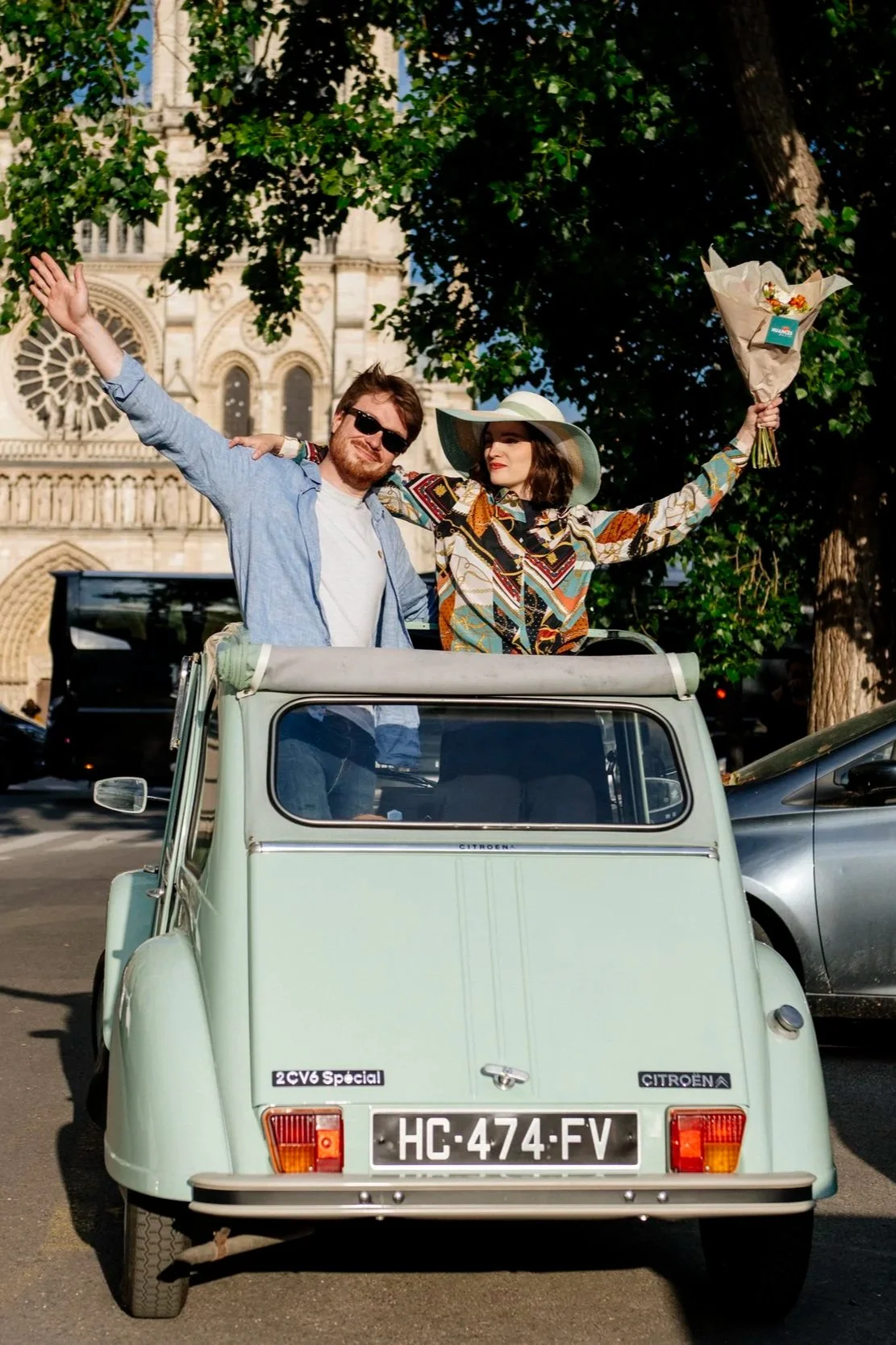 Un couple souriant se tient dans un véhicule ancien à Paris, avec la cathédrale Notre-Dame en arrière-plan. La femme porte un chapeau et une robe colorée, tient un bouquet de fleurs, tandis que l'homme porte des lunettes de soleil et une chemise en j