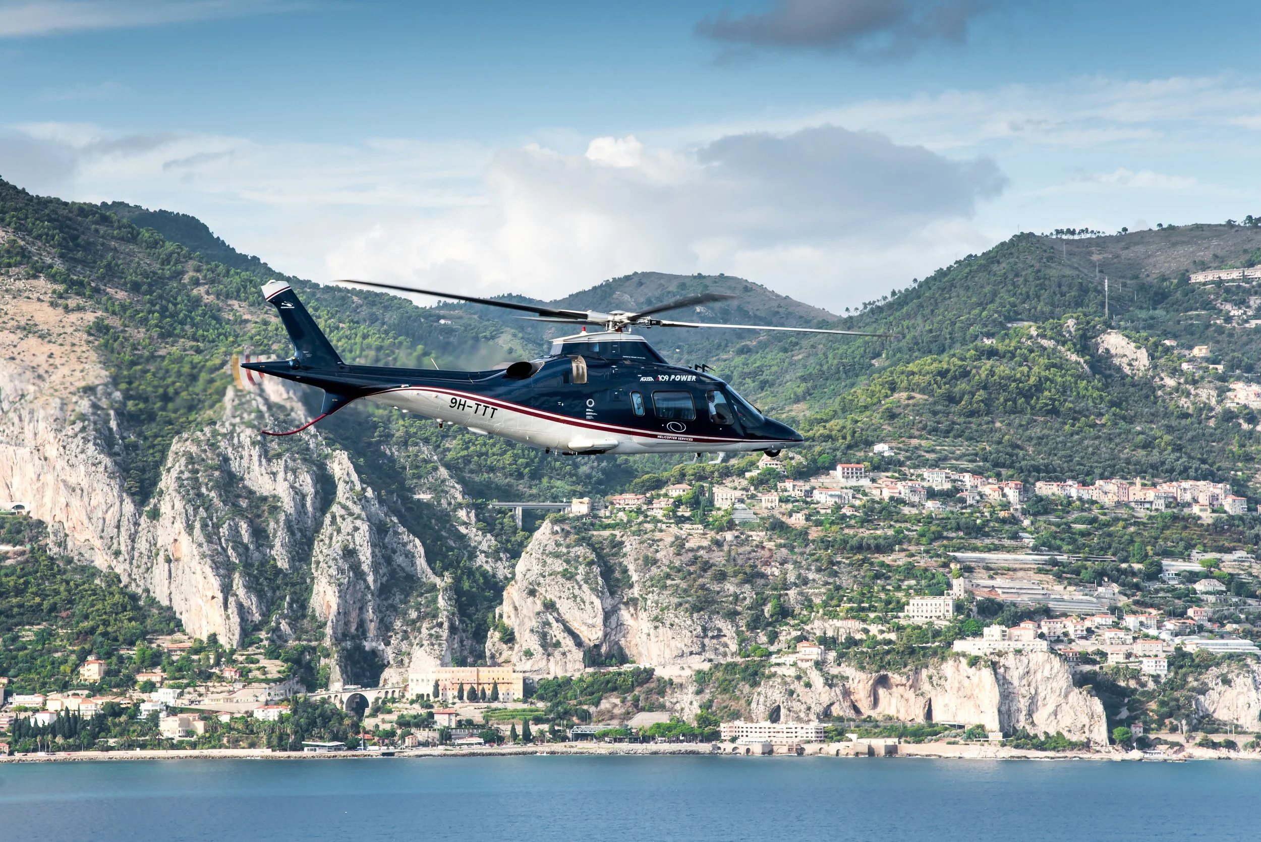 A black helicopter flying over a coastline with hills and buildings in the background.