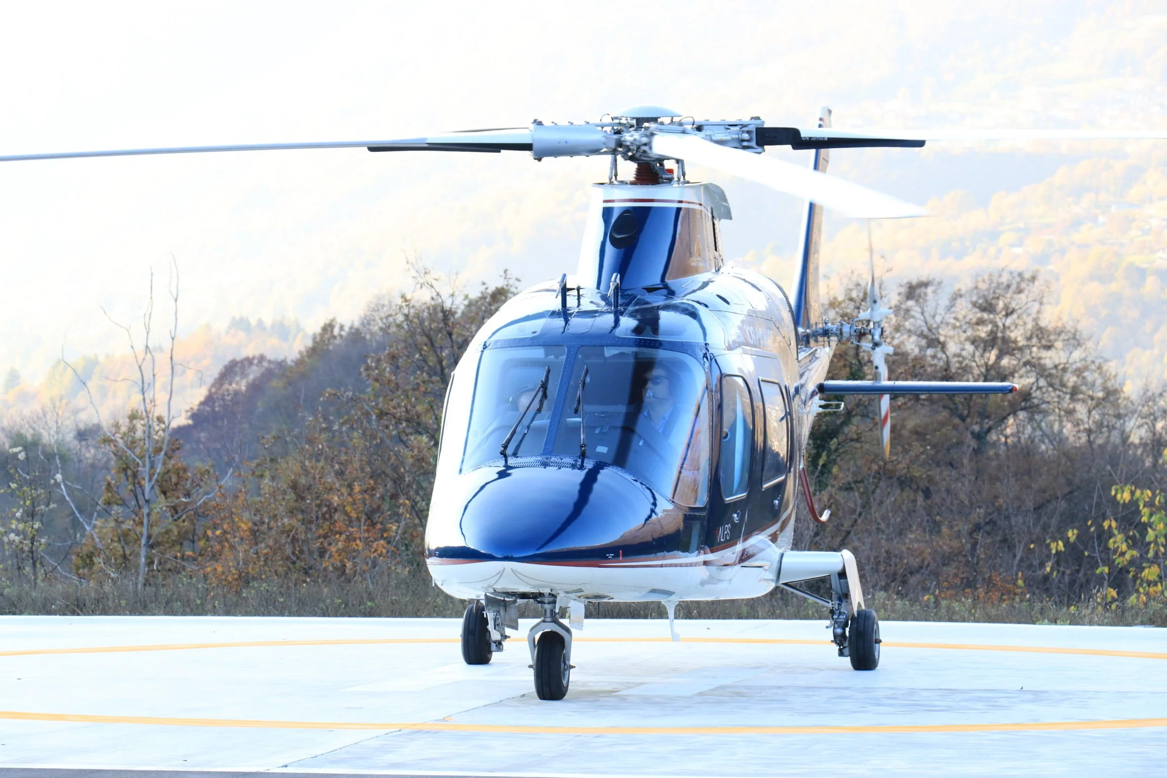 A black and white helicopter on a helipad with trees and mountains in the background.
