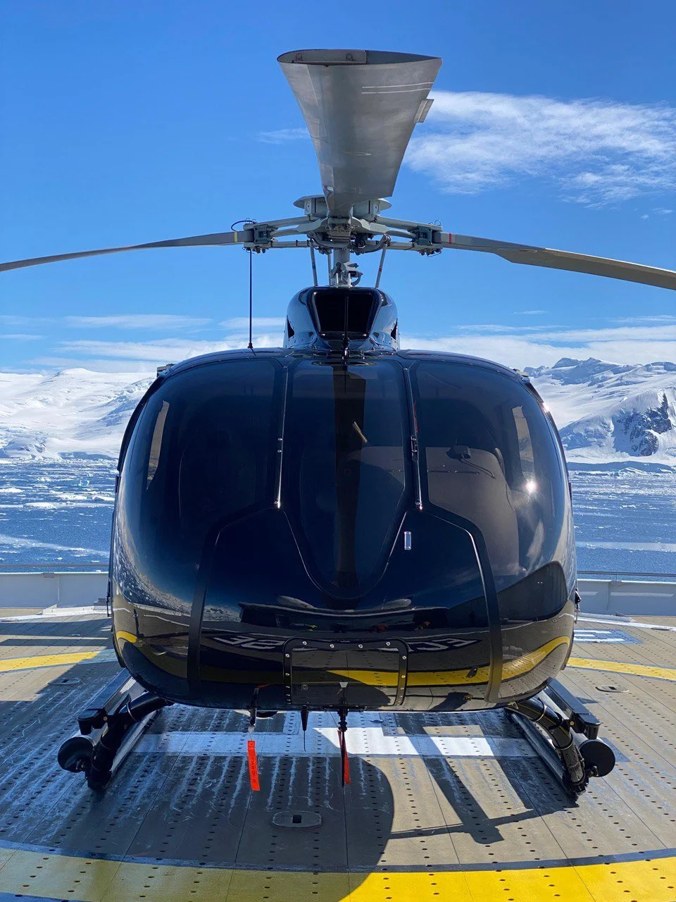 A black helicopter on a helipad with snowy mountains and a clear blue sky in the background.