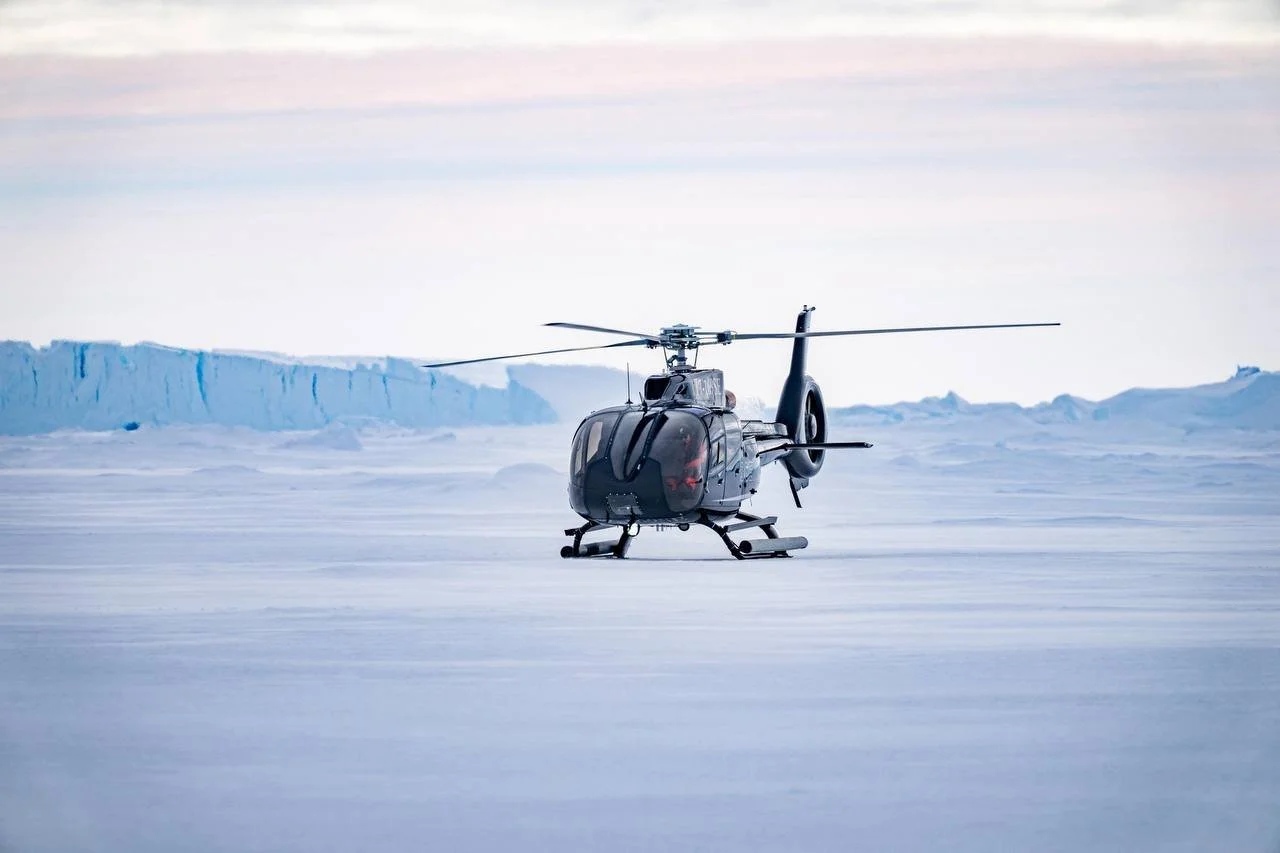 A black helicopter on a snowy, icy landscape with ice cliffs in the background.