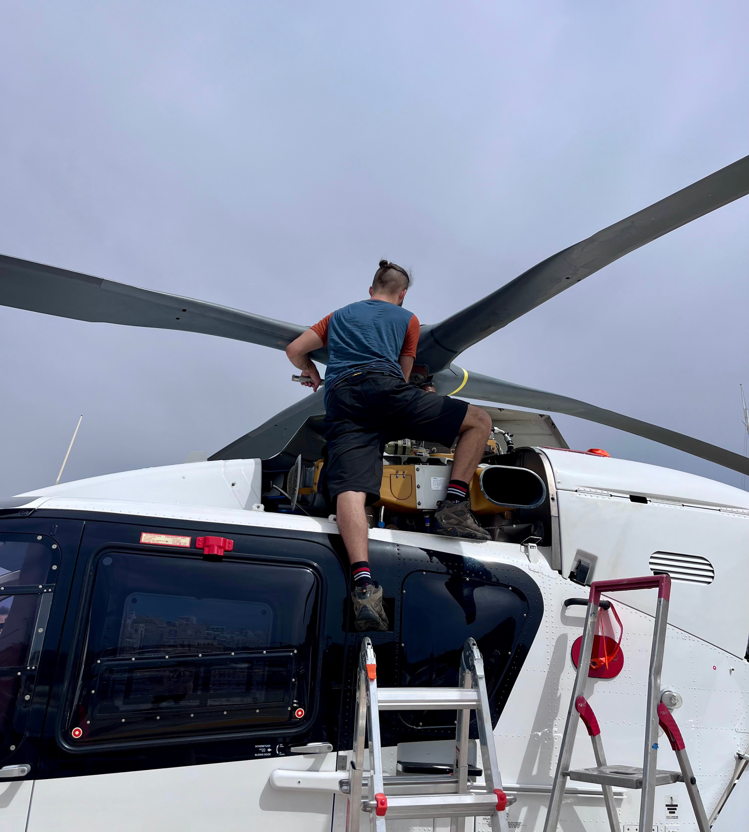 A person working on the engine of a helicopter with access stairs nearby, under a cloudy sky.