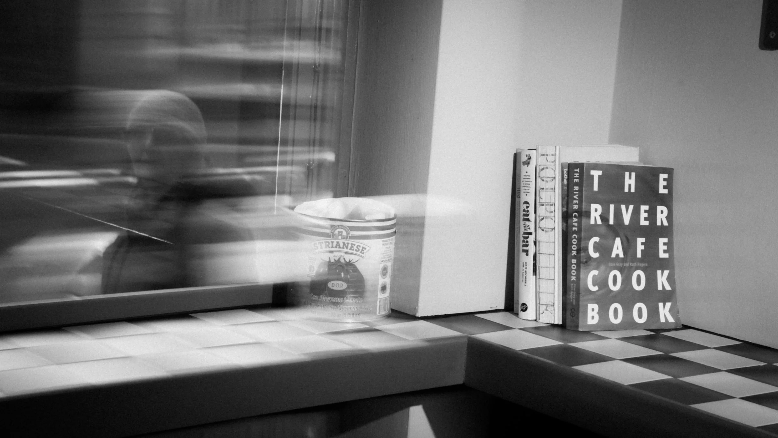 A black and white photograph of a small countertop with a stack of books, a container of Strianese ice cream, and a blurred reflective surface or glass in the foreground.