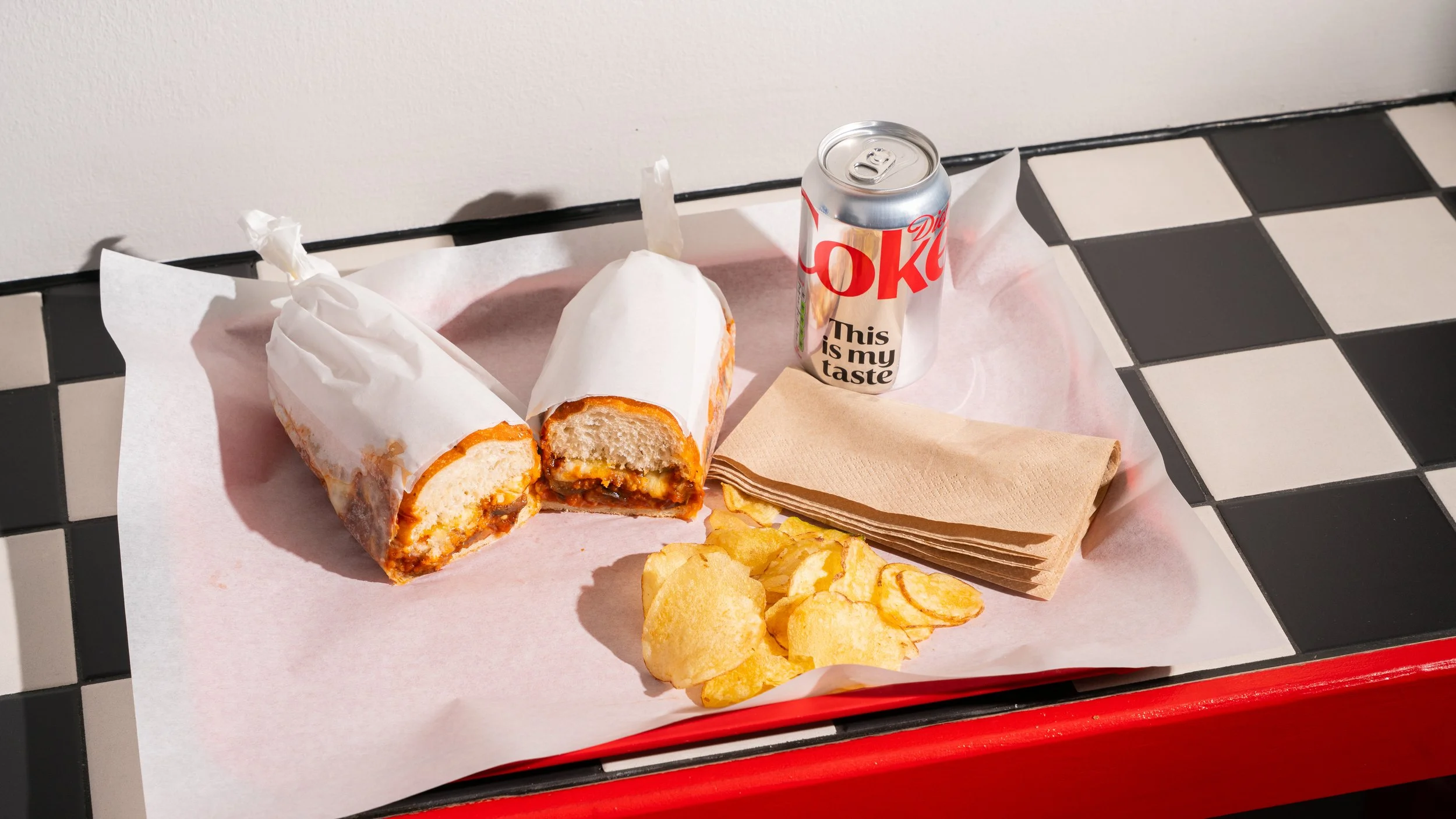 An aubergine ragu sandwich in white paper, a can of Diet Coke, crisps and a stack of napkins on a red tray with pink paper lining, on a black and white checkered table.