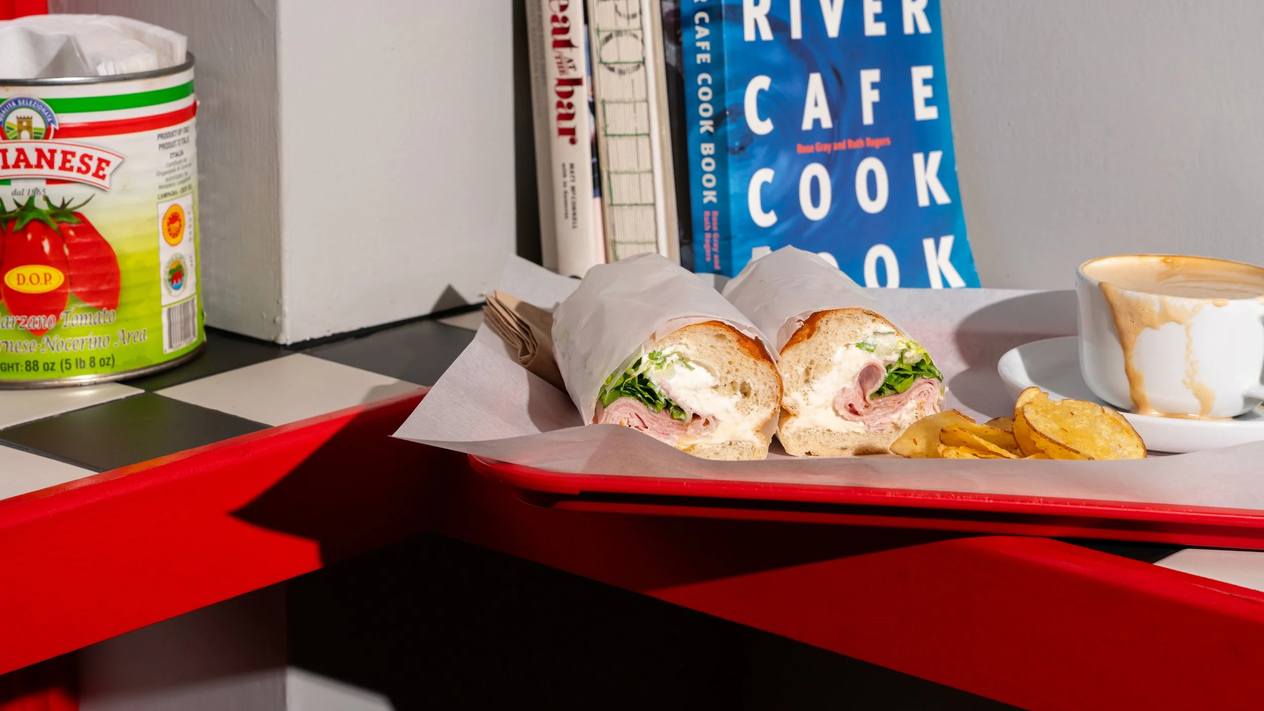 A sandwich cut in half with prosciutto ham, burratta, romaine lettuce & honey served on a red tray with Yorkshire crisps, a cup of coffee with foam, and a book titled 'River Cafe Cook Book' in the background.