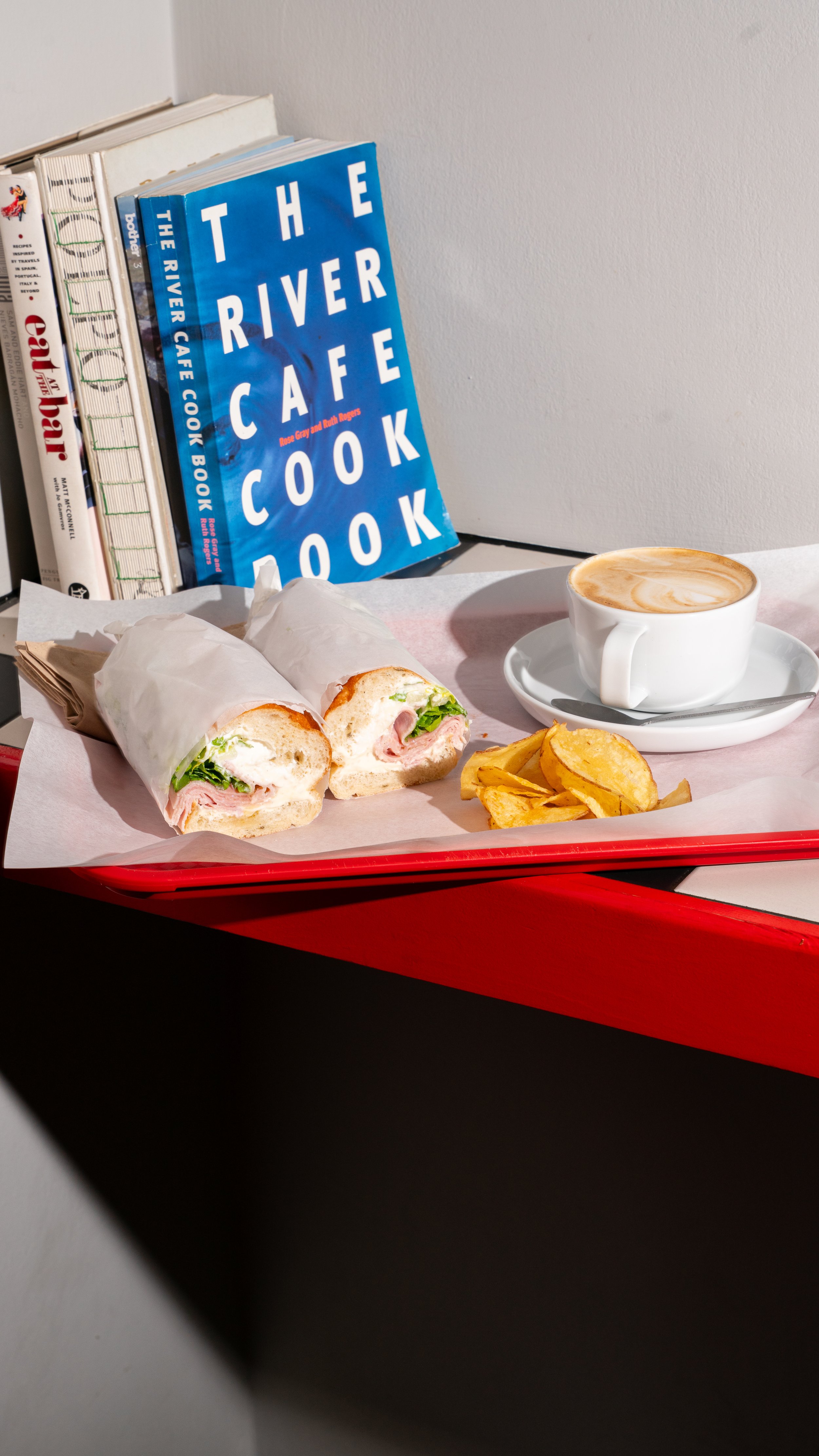 Sandwich with ham, lettuce, and cream, served with potato chips, a cup of coffee with latte art, and a side of potato wedges on a red tray, with cookbooks in the background.