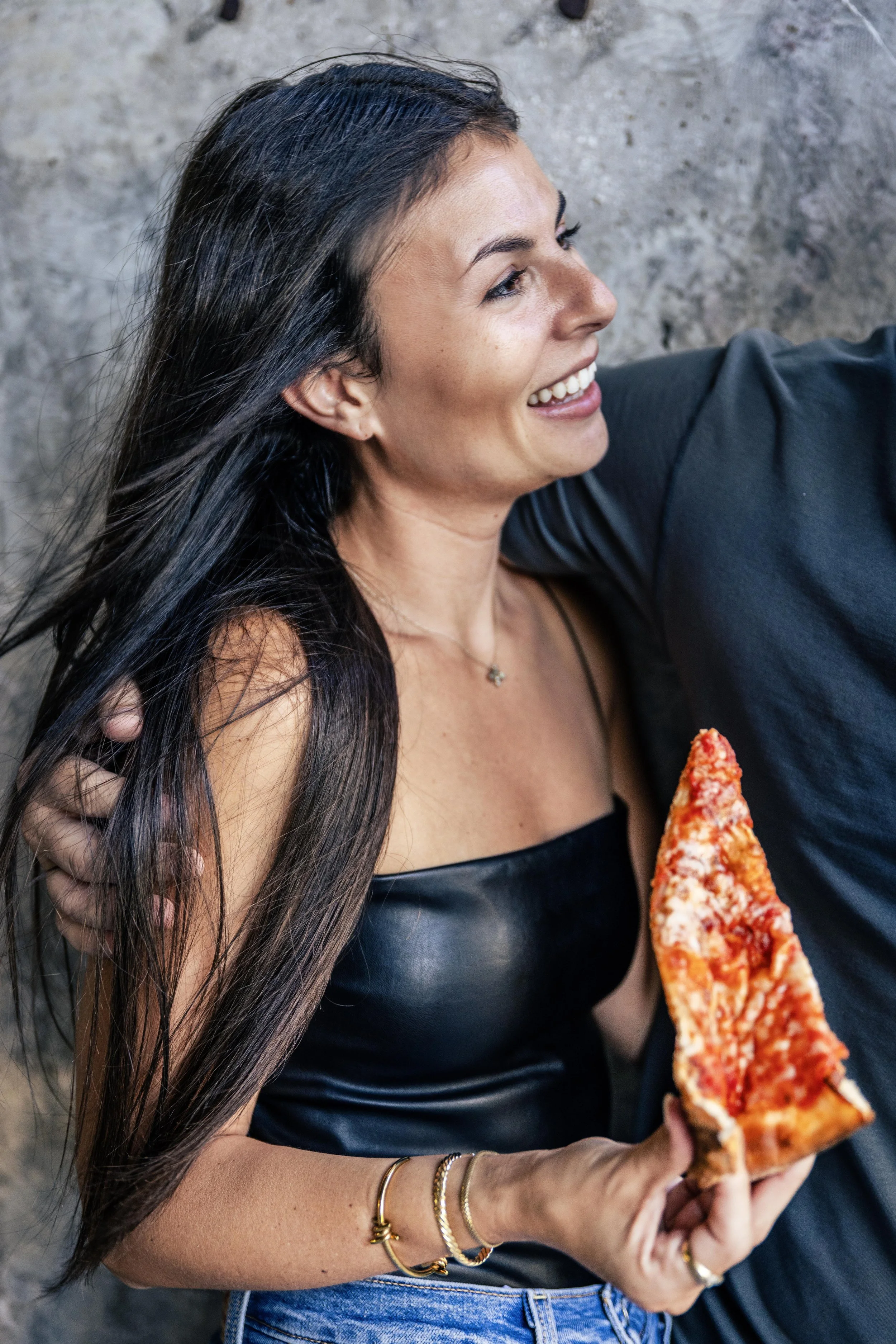 A smiling woman with long dark hair holding a slice of pizza, standing against a stone wall.