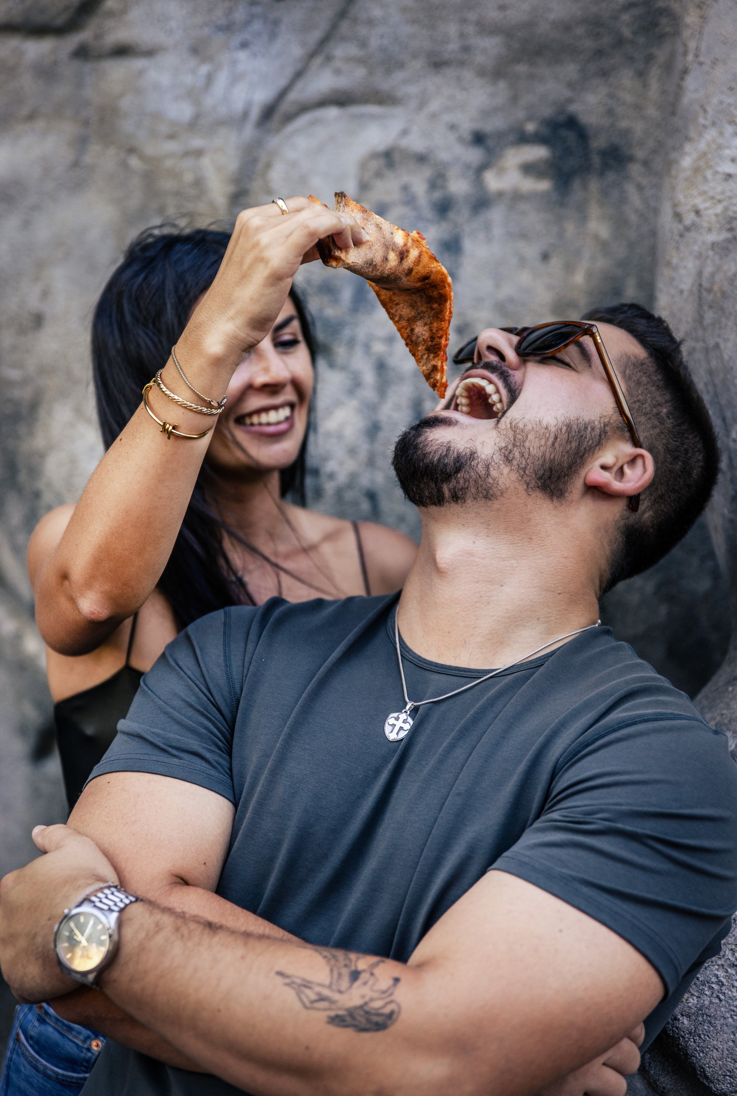 A woman is holding a slice of pizza above a man's mouth while he prepares to bite it. They are standing against a stone wall, smiling and laughing.