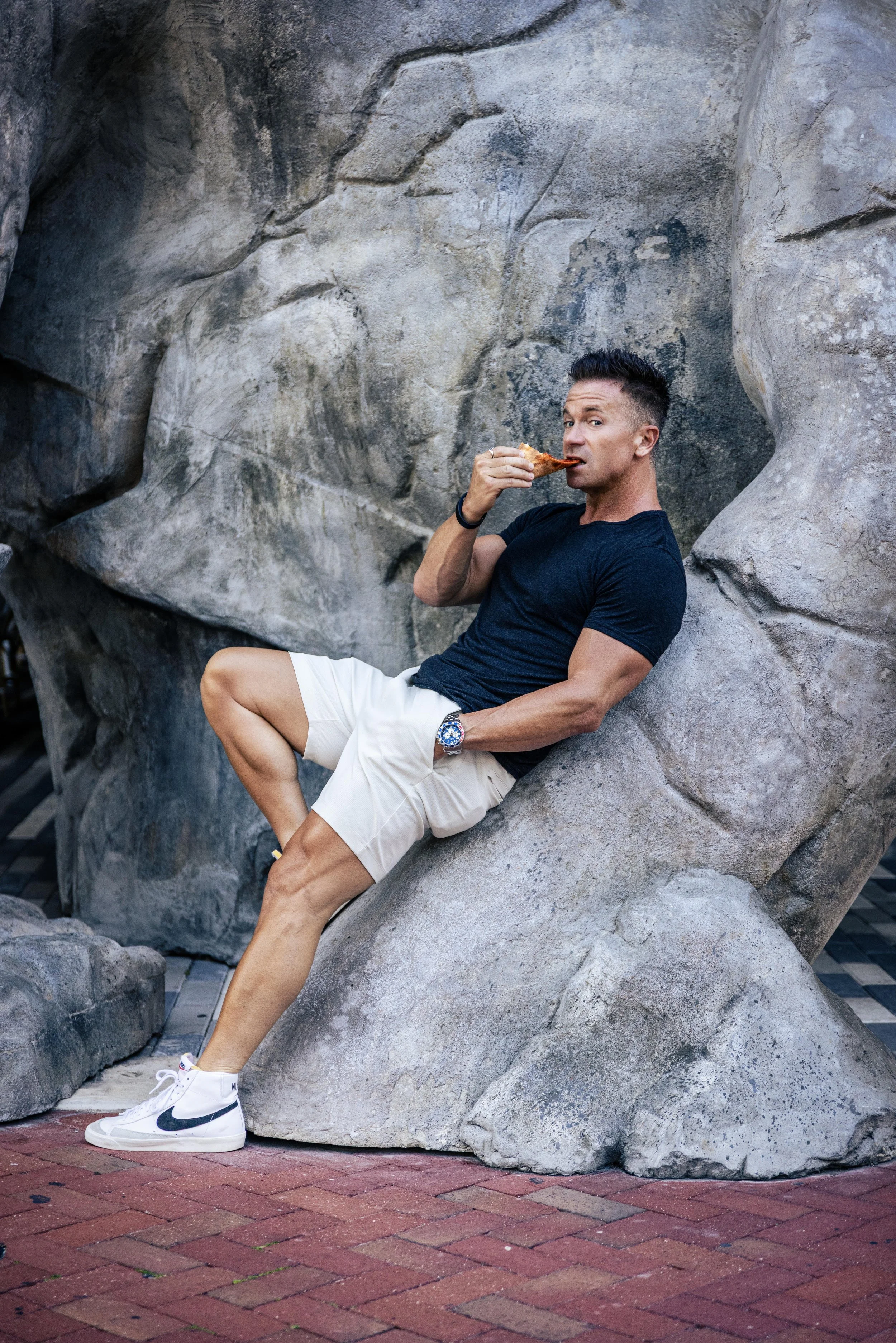 A man sitting on a large rock formation, eating a slice of pizza, wearing a black t-shirt, white shorts, and white sneakers, with a city sidewalk in the background.