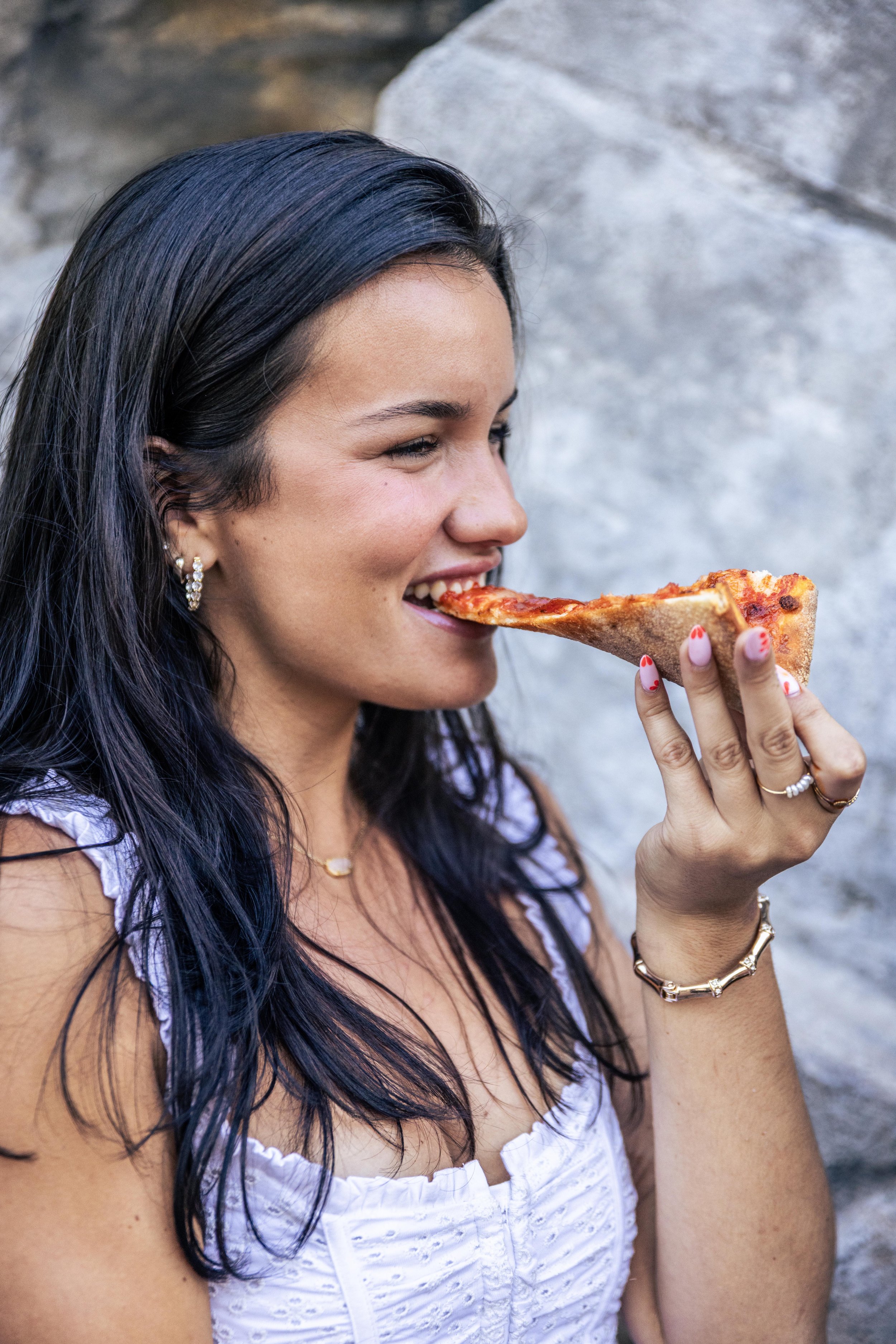 A woman with dark hair and jewelry is eating a slice of pepperoni pizza outdoors.