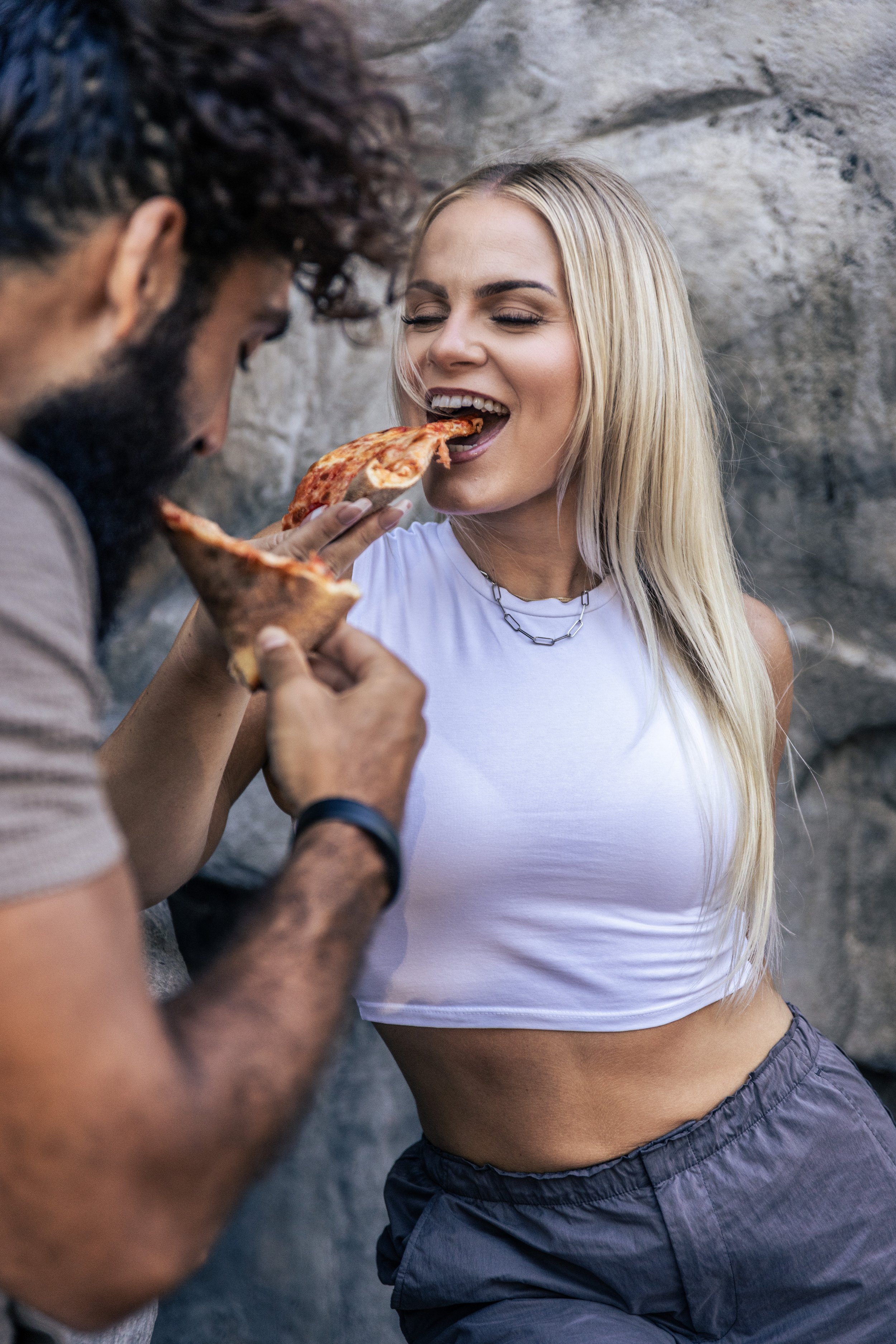 A woman with long blonde hair in a white tank top and gray shorts is smiling as a man with curly hair and beard feeds her a slice of pizza outdoors against a rocky background.
