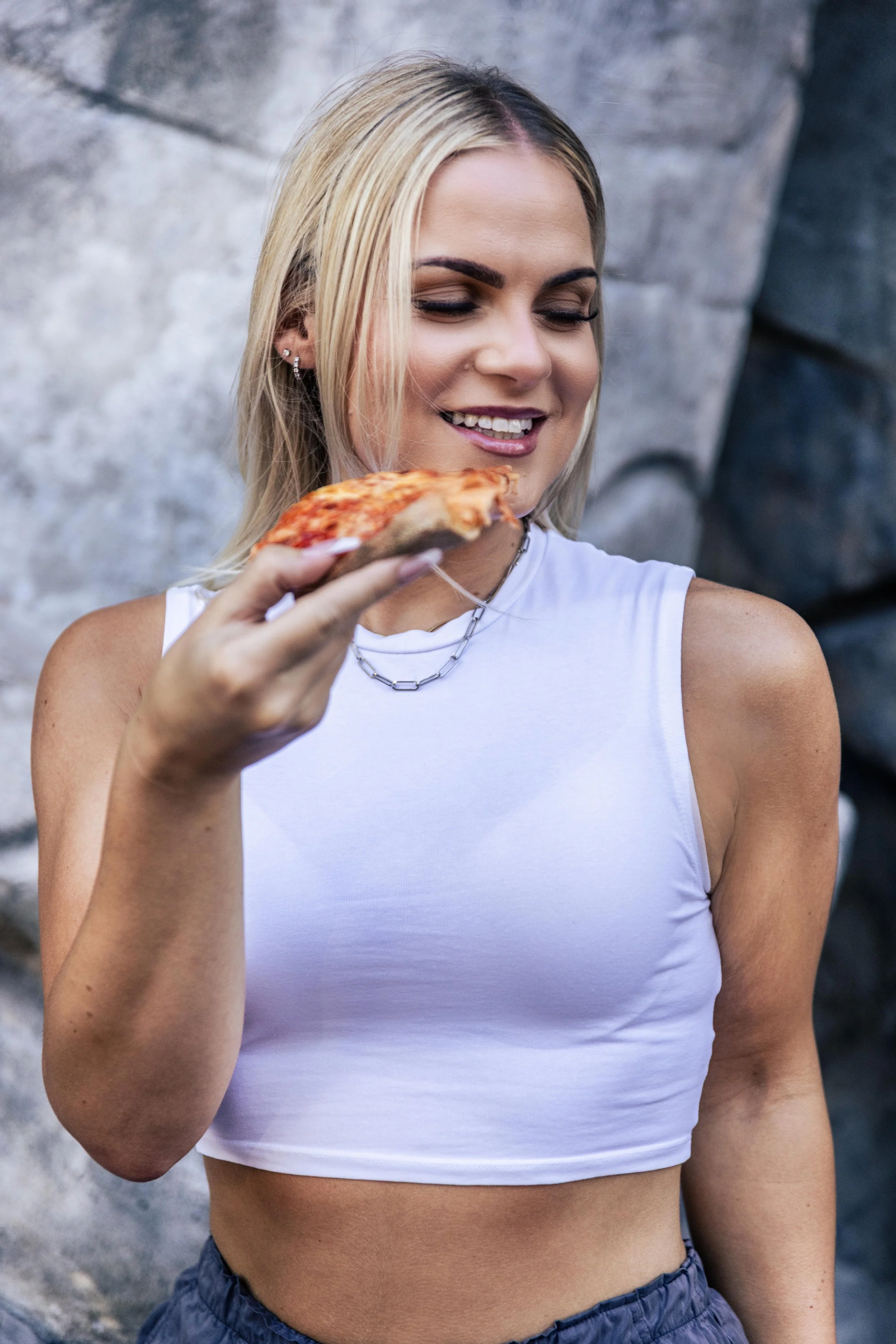 A woman holding a slice of pizza, smiling and looking at it, standing outdoors against a stone wall.