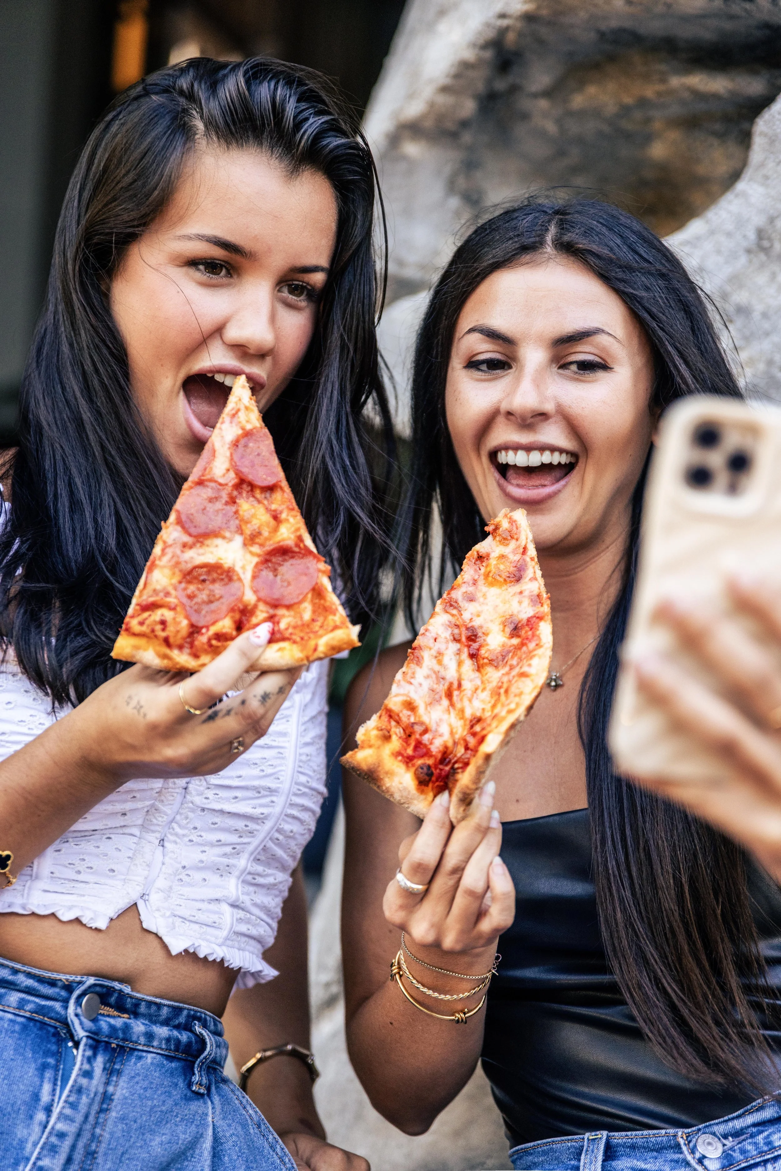 Two women smiling and taking a selfie while holding slices of pepperoni pizza.