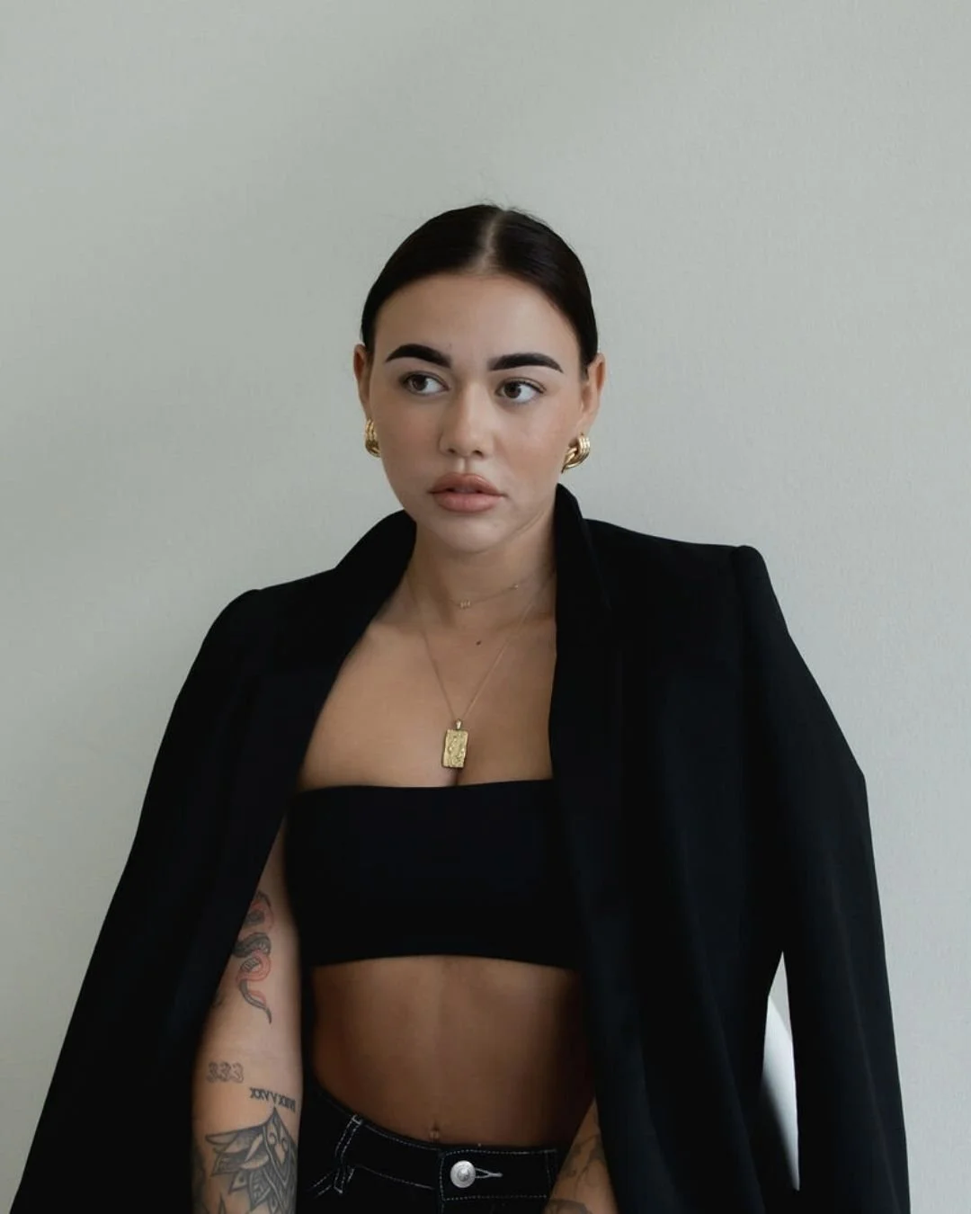 A young woman with dark hair pulled back, wearing gold jewelry, a black cropped top, and a black blazer. She has tattoos on her left arm and is standing against a plain off-white wall.