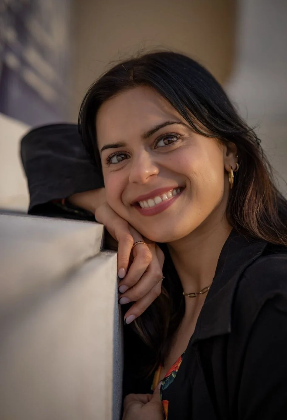 A woman with dark hair and hoop earrings smiling and resting her head on her hand, leaning on a ledge or table.