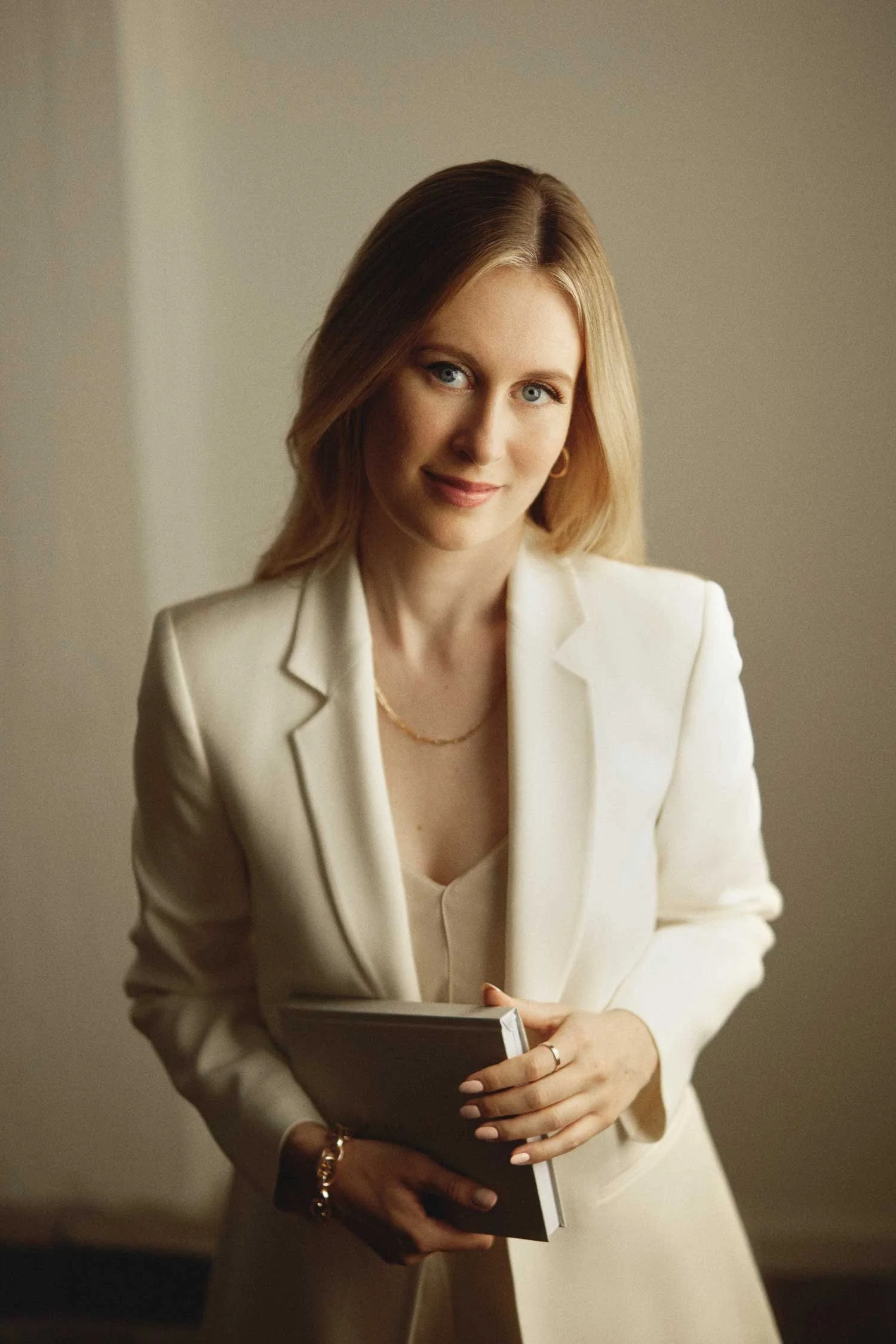 A woman with blonde hair, wearing a white blazer and holding a notebook, standing indoors and smiling softly at the camera.