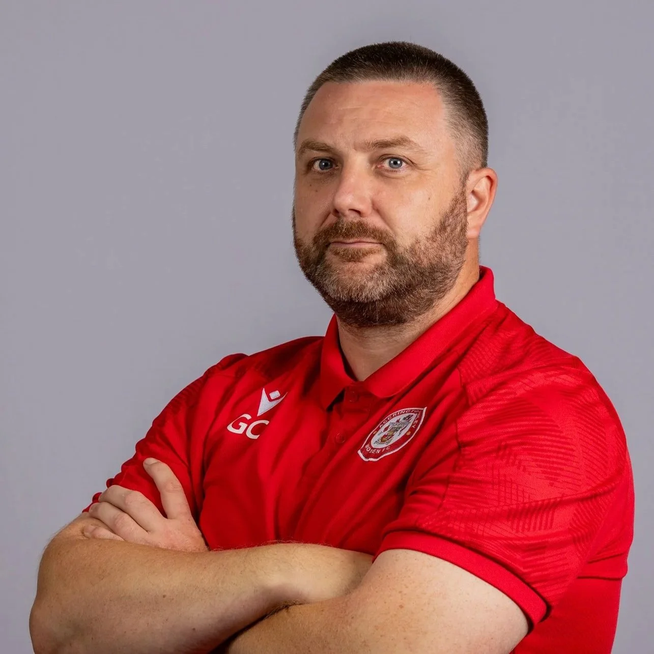 A man with a beard and short hair wearing a black sports jacket with a logo of Accrington Stanley Women FC, standing in front of a backdrop with logos of IMEP, Sund, and Accrington Stanley Women FC.