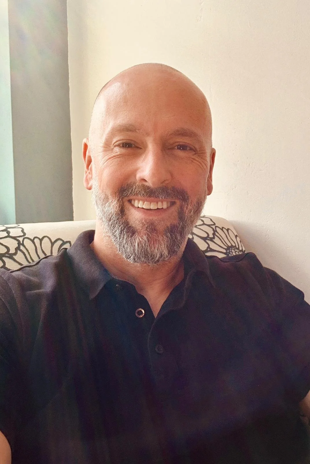 A smiling man with a beard and bald head, wearing a black polo shirt, sitting on a couch with a floral pillow in the background.