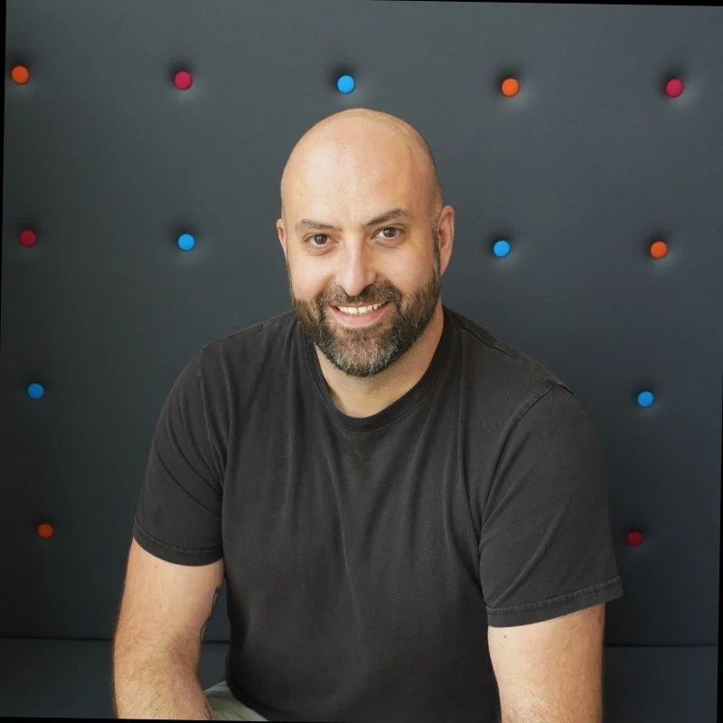 A smiling man with a beard and bald head wearing a black t-shirt, sitting against a dark wall with colorful small round decorations.