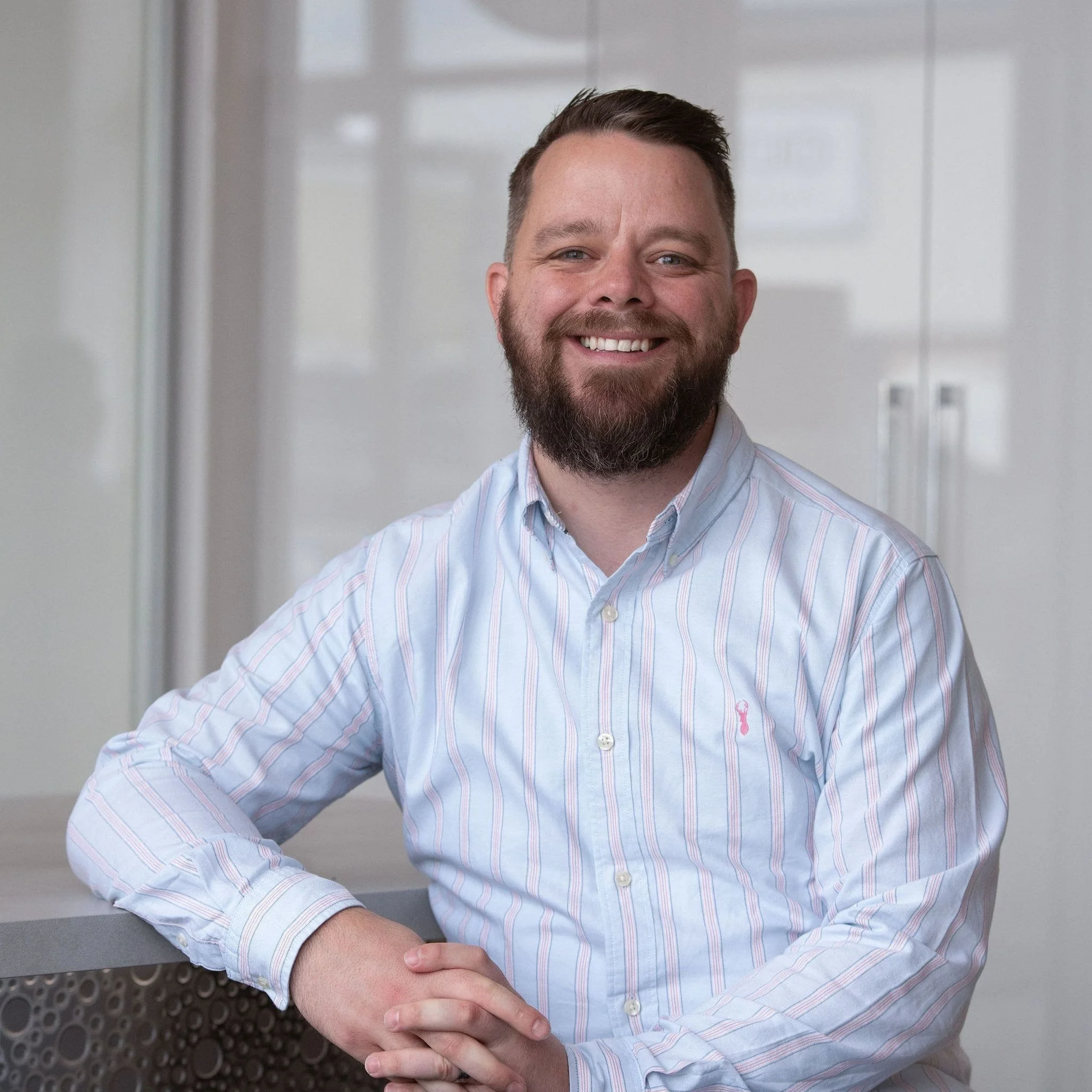 A smiling man with a beard and short hair, wearing a light blue, striped button-up shirt, standing in an office environment.