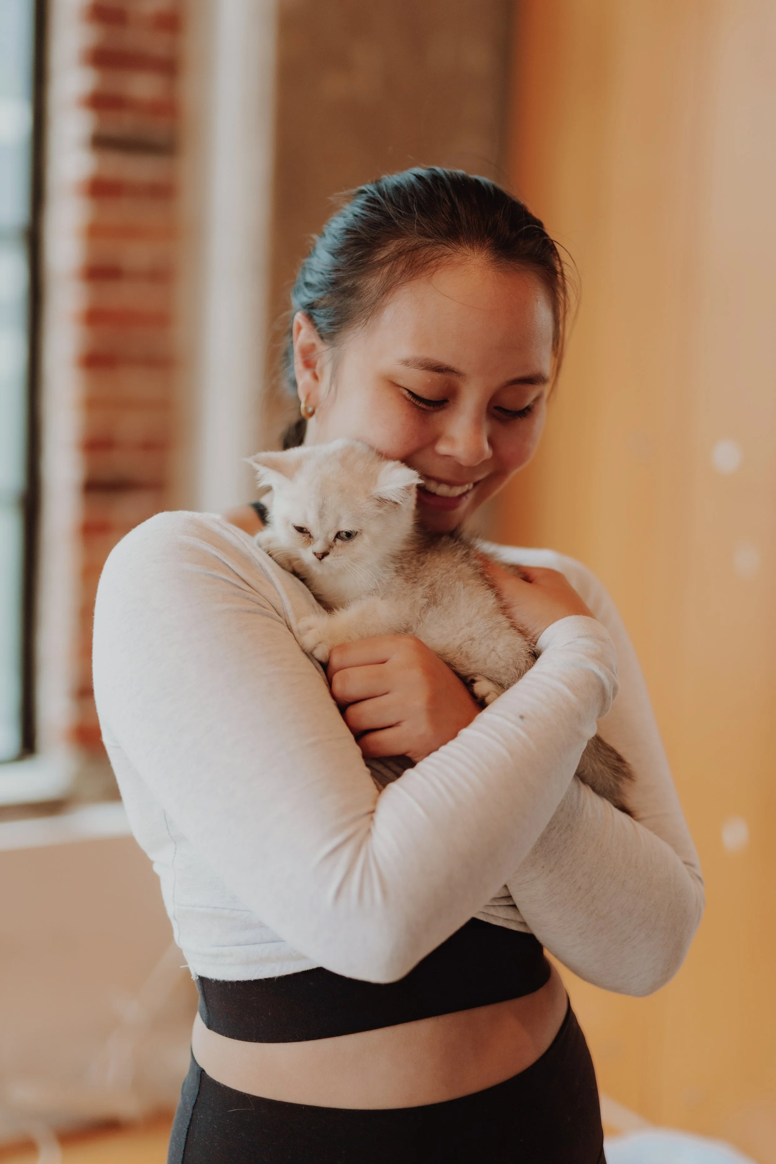 A woman is smiling and hugging a gray and white kitten indoors.
