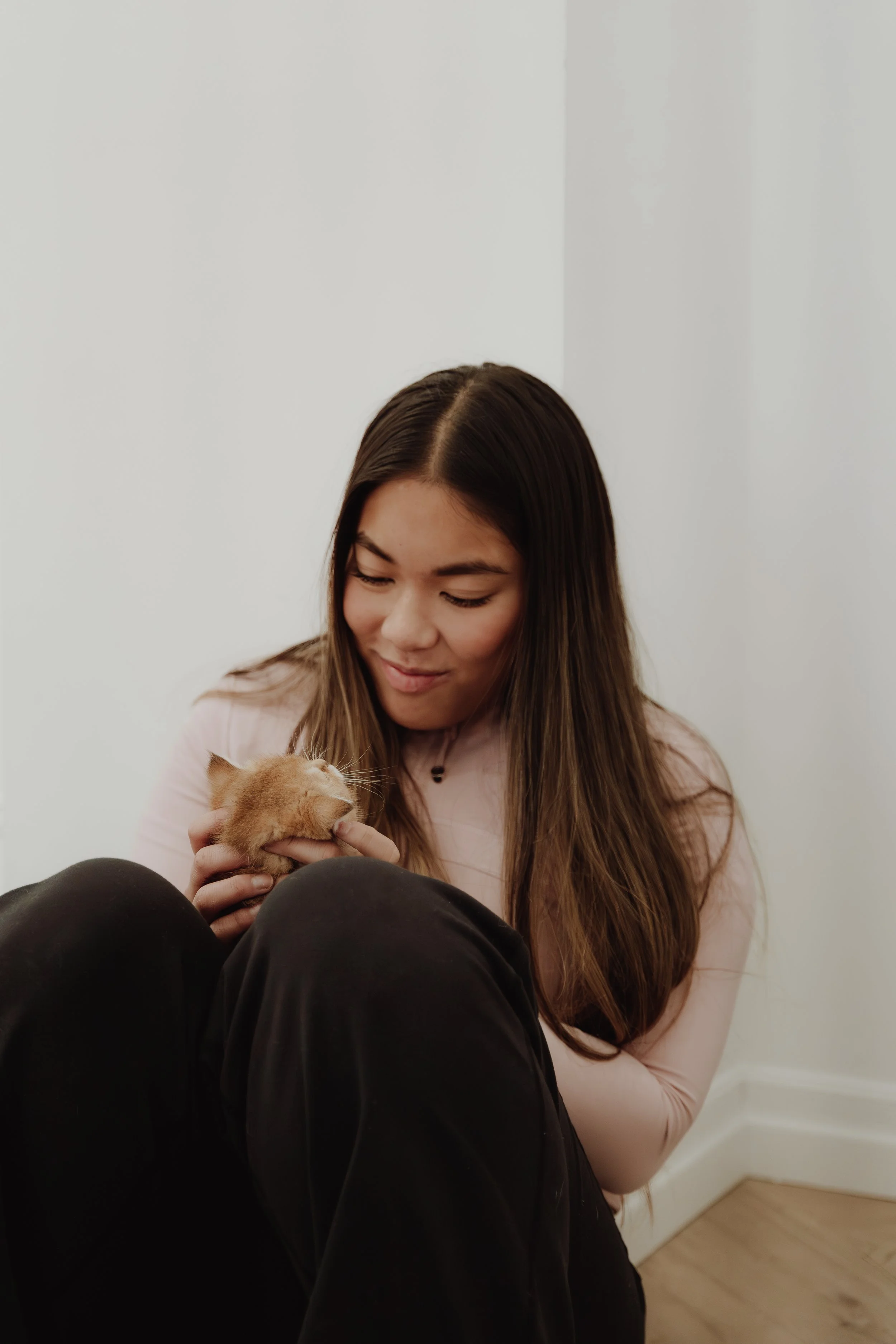 A woman is sitting on the floor holding a ginger kitten near her face with a gentle smile, indoors with a plain white wall in the background.