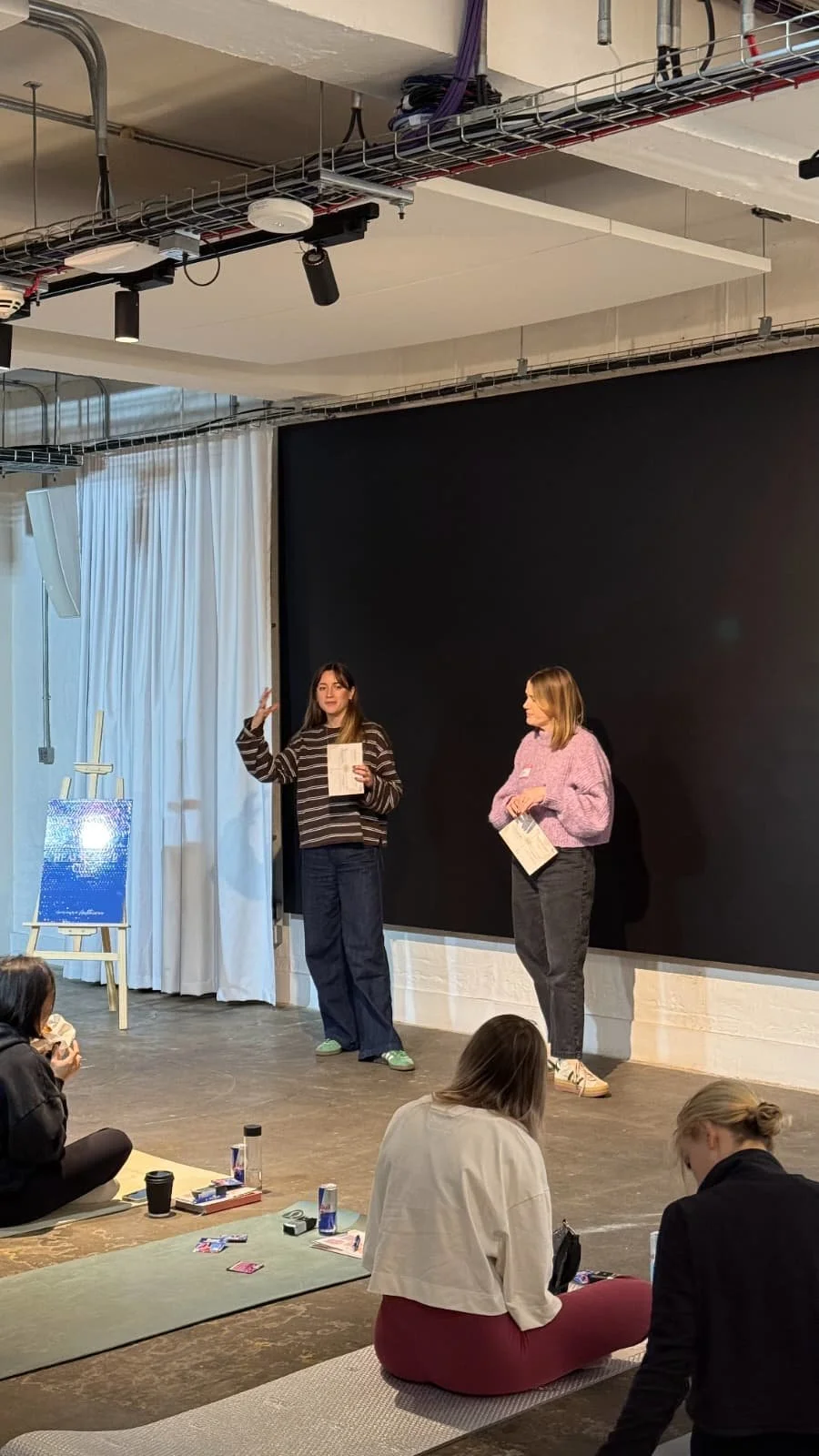 Two women are standing on a stage, speaking to an audience, holding papers. A painting on an easel and white curtains are visible behind them. Audience members are sitting on mats on the floor, some holding drinks.