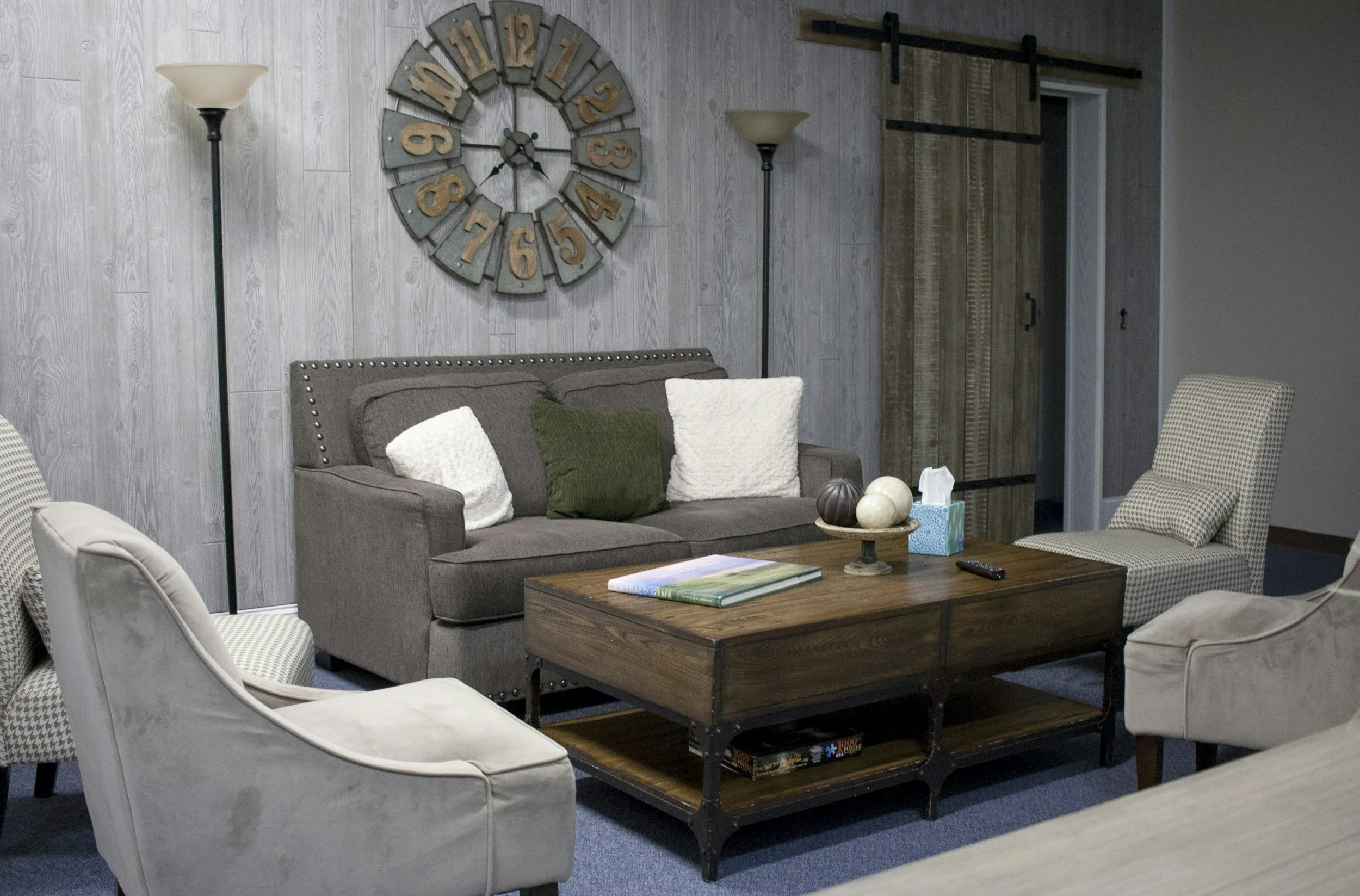 A cozy living room with a gray upholstered sofa with three pillows, a wooden coffee table with decorative balls and a tissue box, surrounded by armchairs, and a large wall clock with numbers in a circular pattern mounted on a wood-paneled wall. There are two tall floor lamps on either side of the sofa and a sliding barn door to the right.