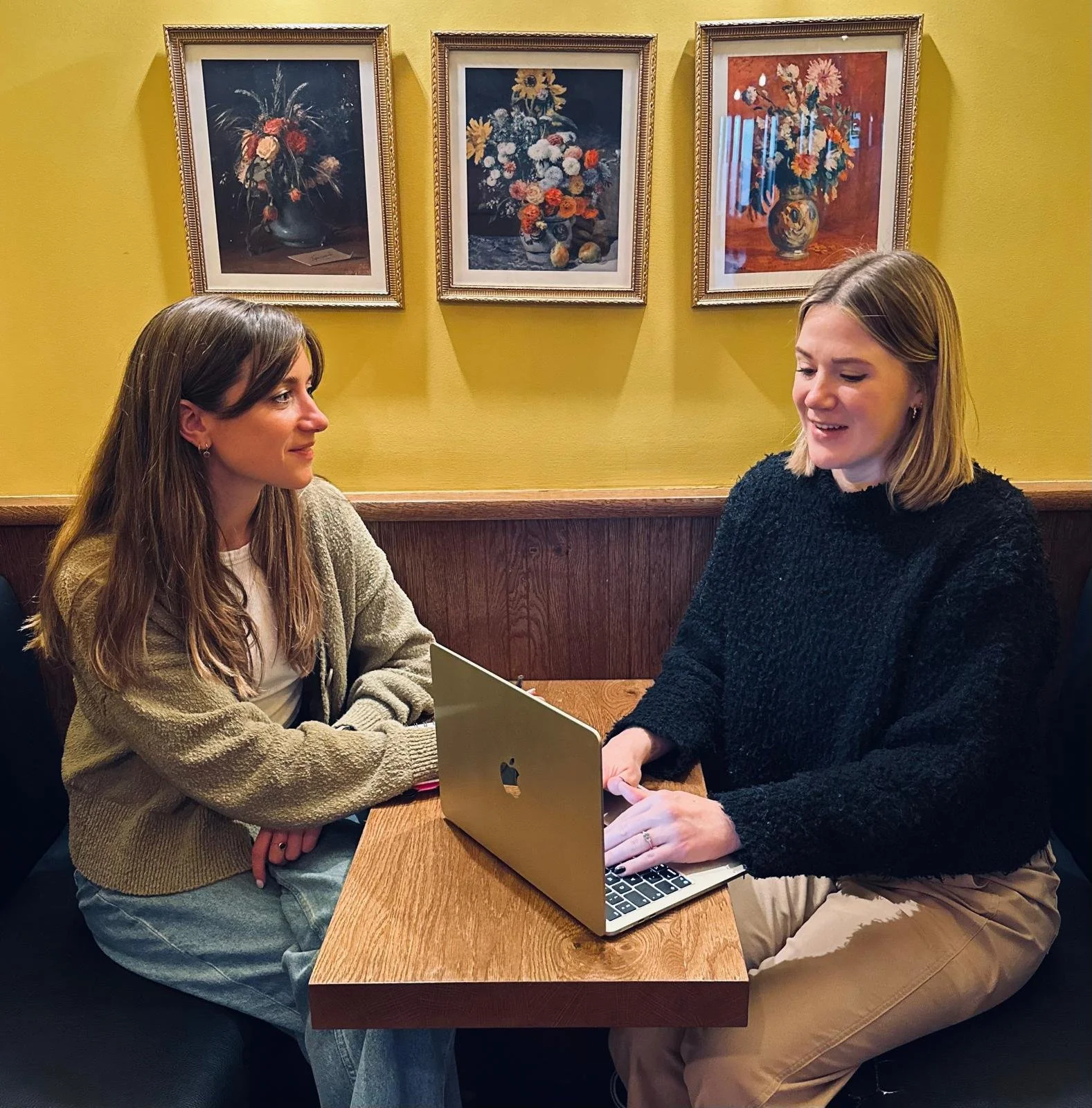 Two women sit at a table facing each other. One is on a laptop.