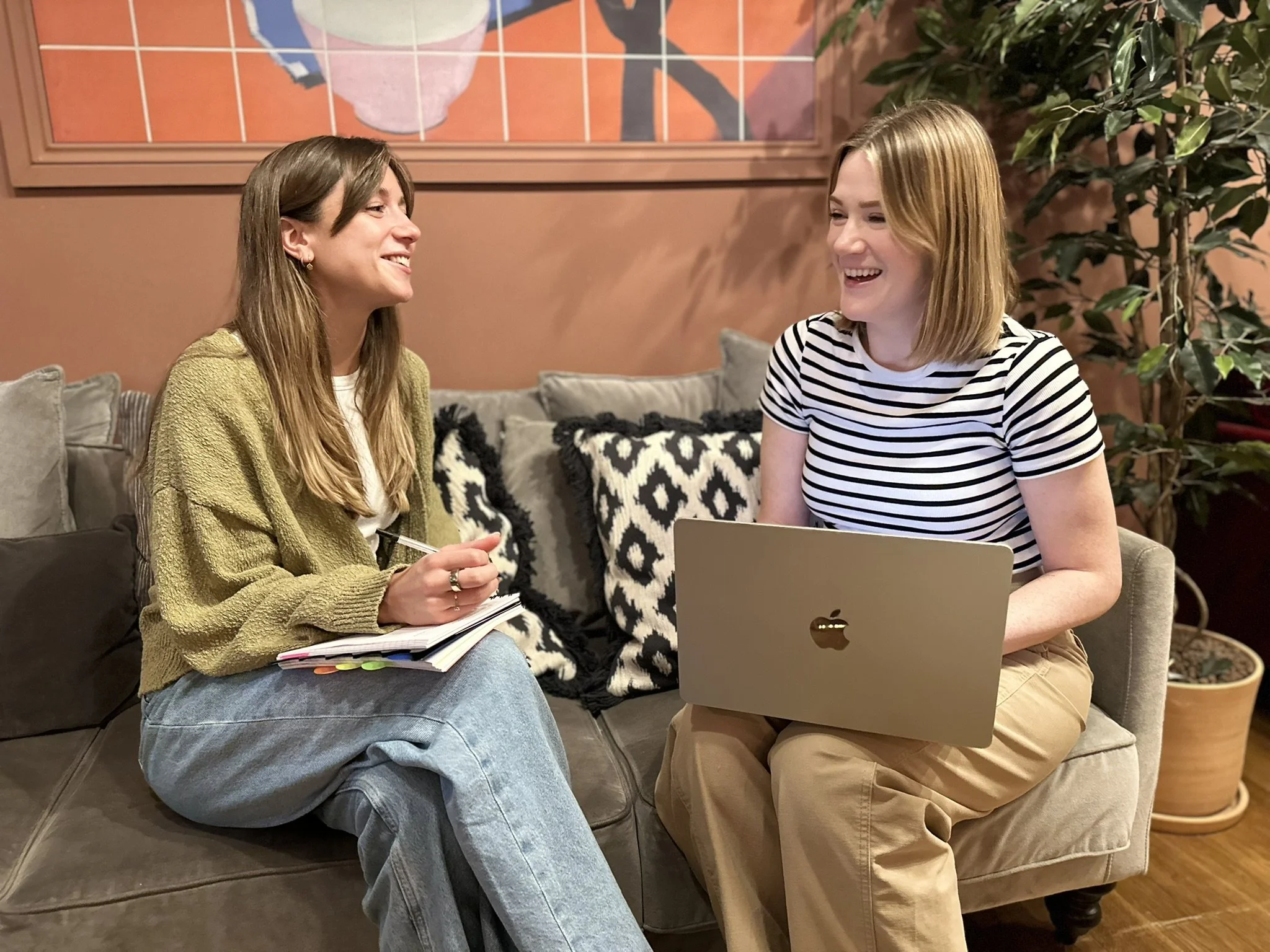 Two women sitting on a grey sofa with patterned cushions, smiling and talking; one holding a notepad, the other using a laptop.