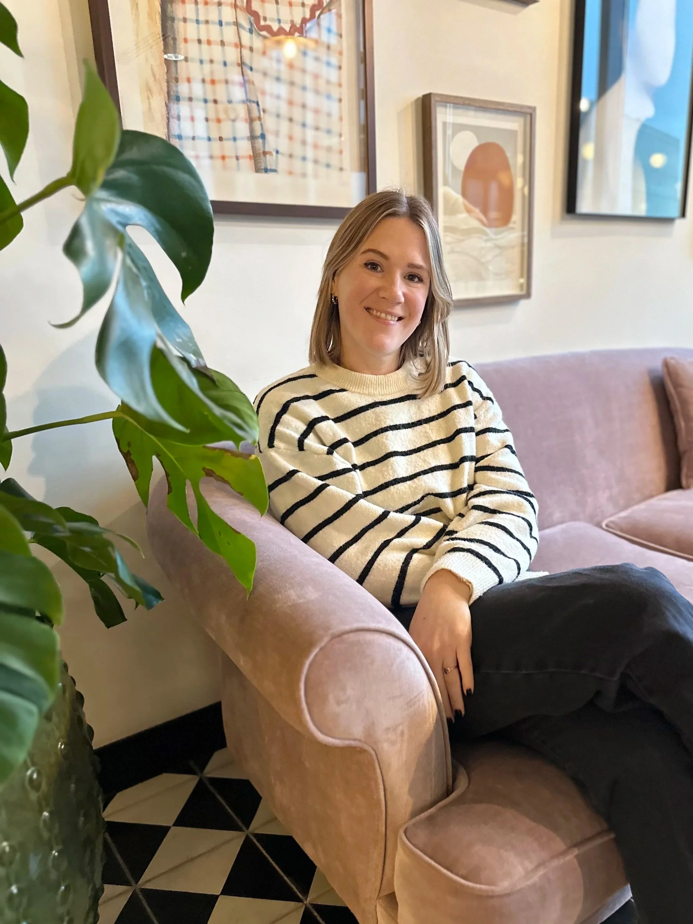 A smiling woman with shoulder-length blonde hair, wearing a cream and black striped sweater, sitting on a pink armchair in a cozy, art-decorated room. There is a green plant in the foreground and framed artwork on the wall behind her.