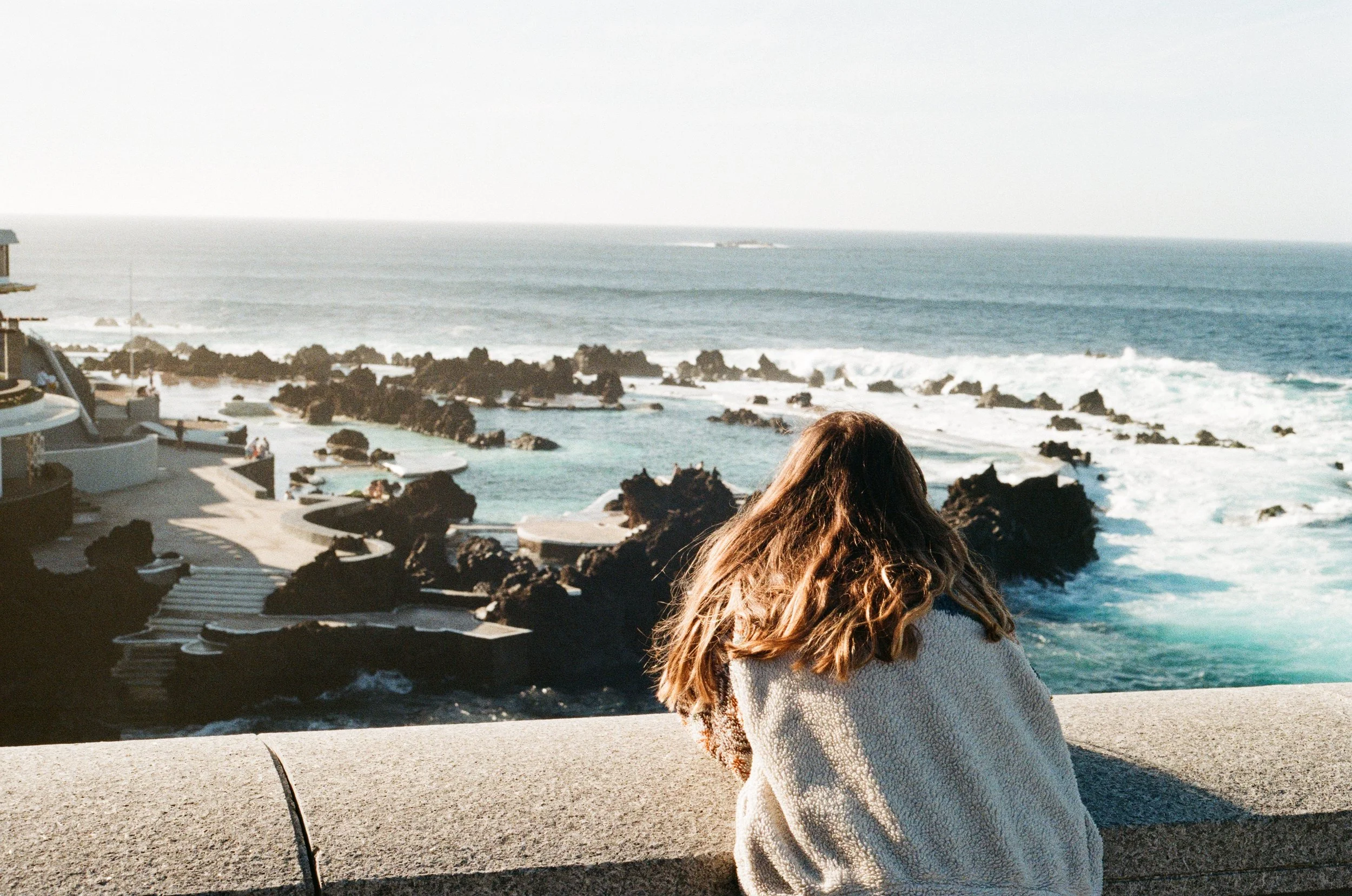 Woman with long brown hair in a fleece leaning on a wall, looking out over a rocky ocean.