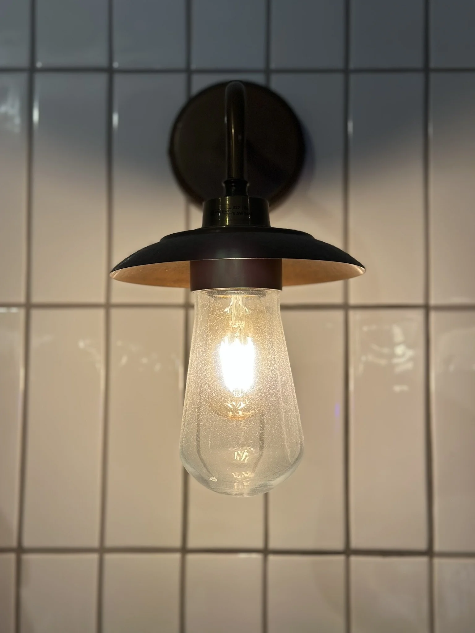 An exposed lightbulb with a shallow lampshade pictured against white tiled wall.