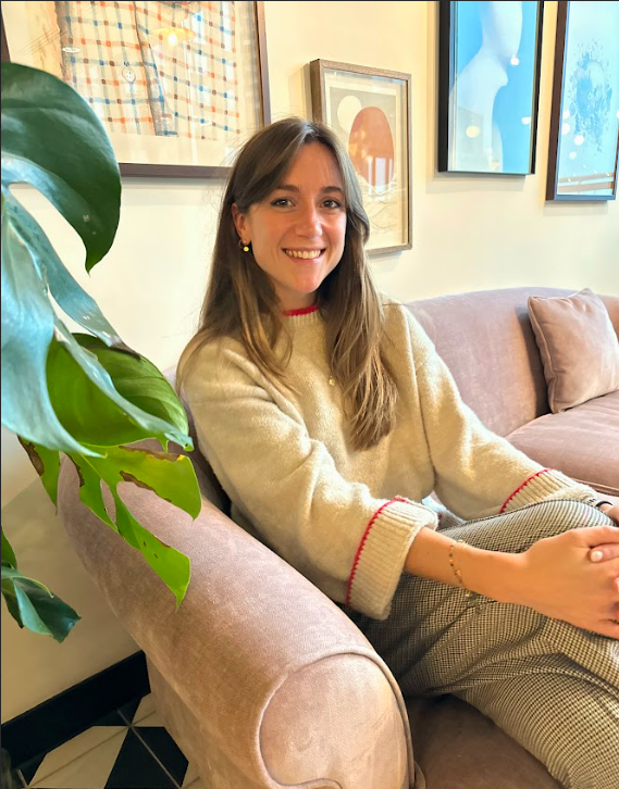A woman with long brown hair and a bright smile sitting on a pink velvet sofa in a well-decorated room with framed artwork on the wall and a large green plant nearby.