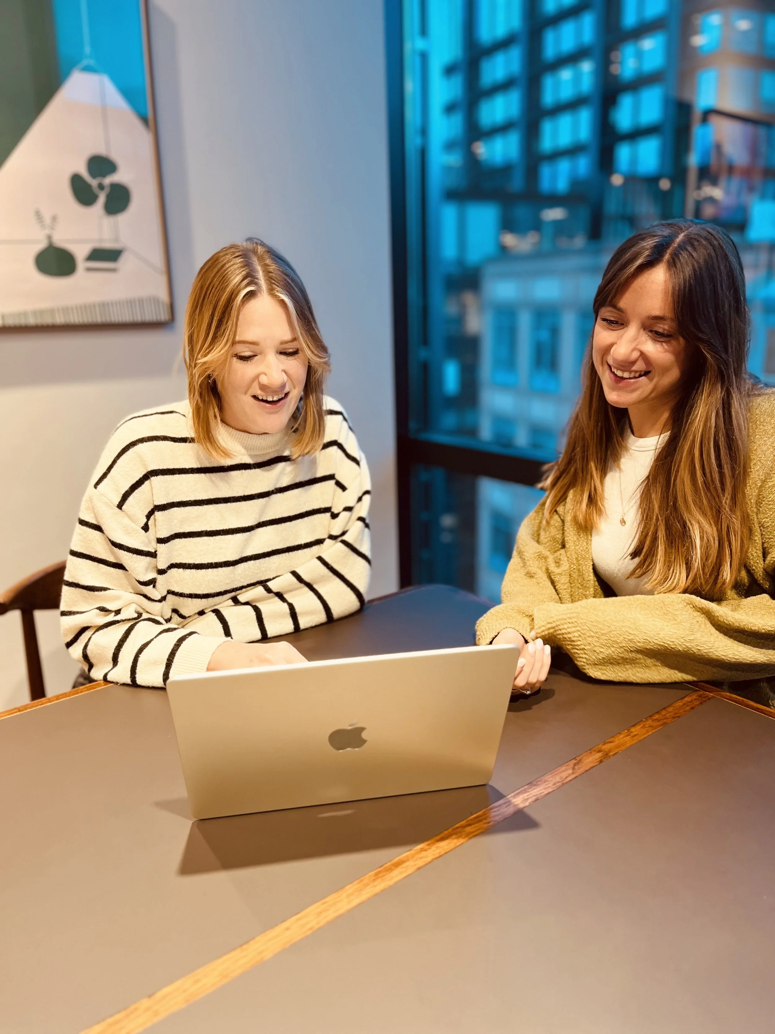 Two young women sitting at a table looking at a laptop, smiling, in a modern room with large windows and a city view.