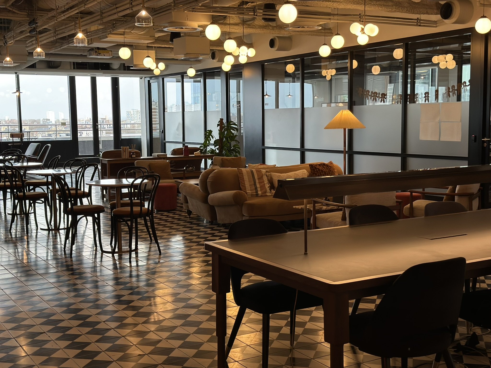Cafe-style cowering space with black and white tiled flooring, tables, sofas, plants and frosted glass windows.
