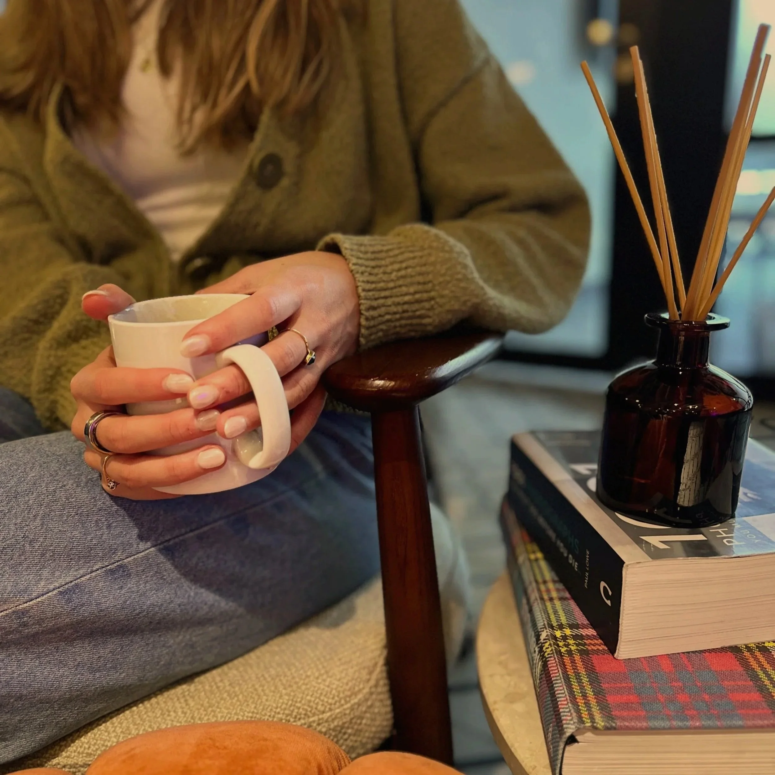 Person in blue jeans and a green cardigan holding a coffee mug, sitting beside a table with books and a reed diffuser.