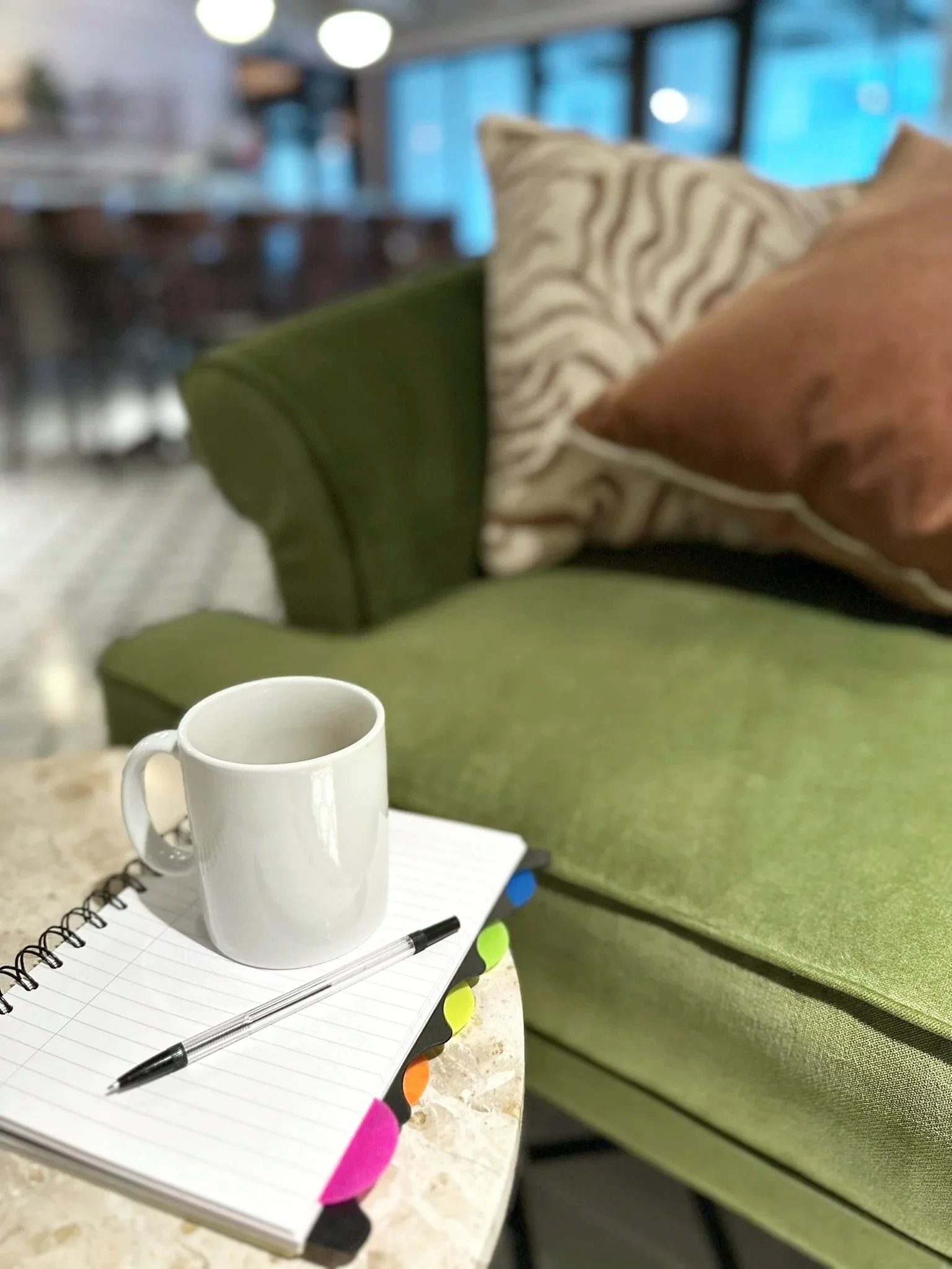 A green velvet sofa with cushions. A marbled side table sits in front of the sofa with a coffee cup, note pad and pen.