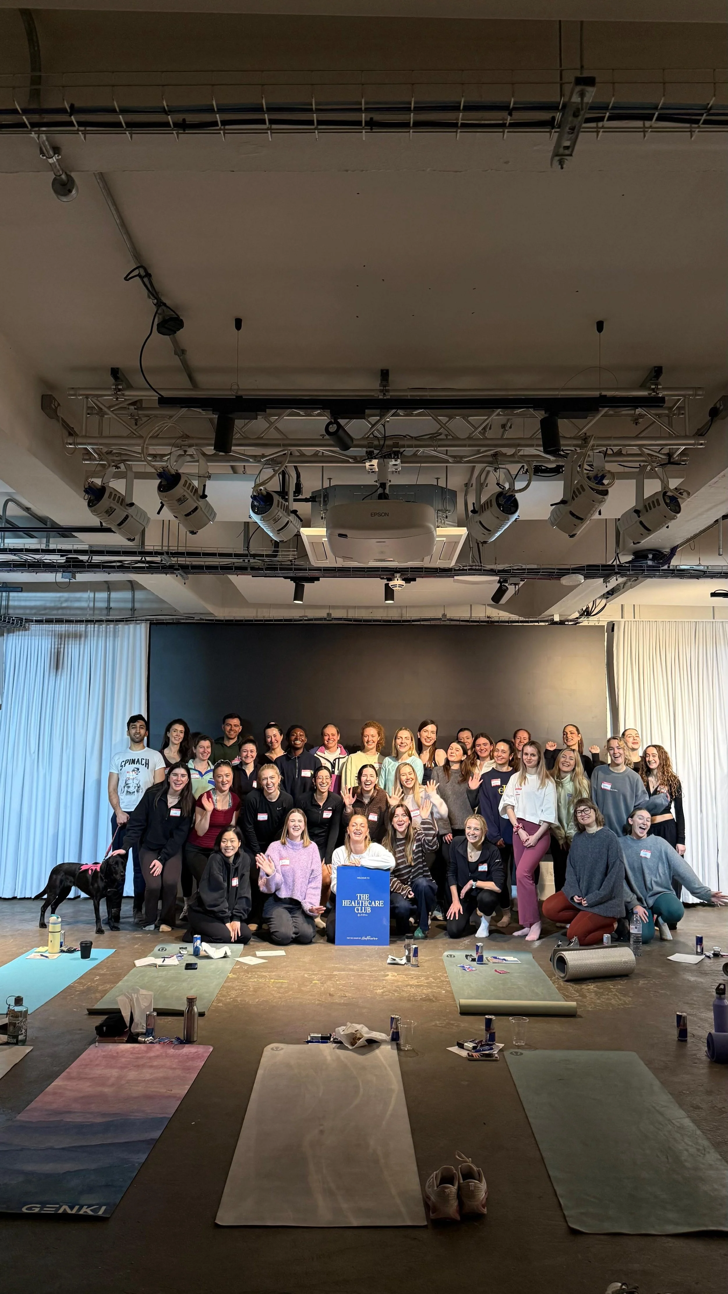A large group of people gathered on a stage, posing for a photo in a studio or event space, with yoga mats and personal belongings on the floor in front of them.