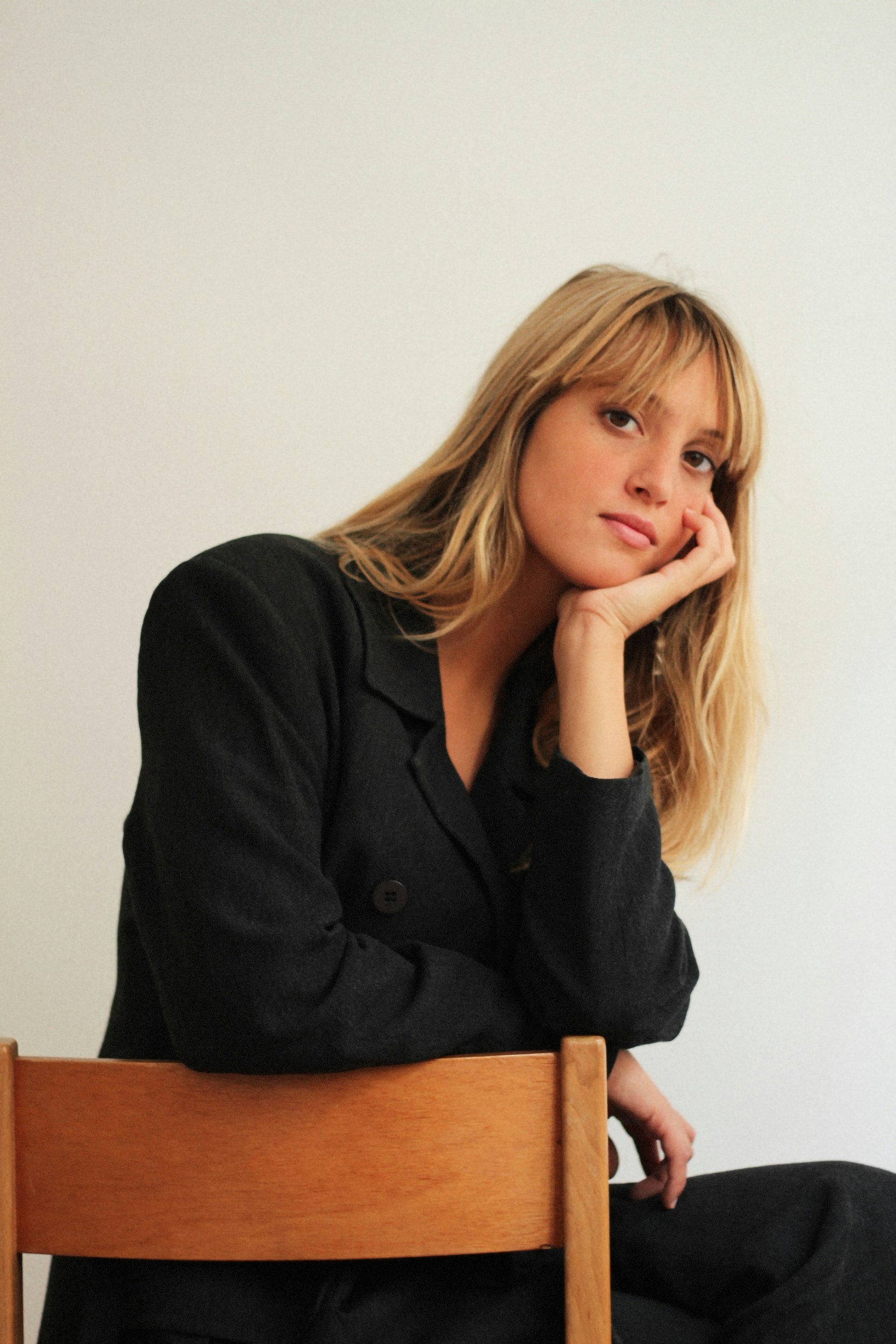 Ung kvinna med långt blont hår, sittande på en stol, med hand under hakan, klädd i svart jacka, mot en vit vägg.