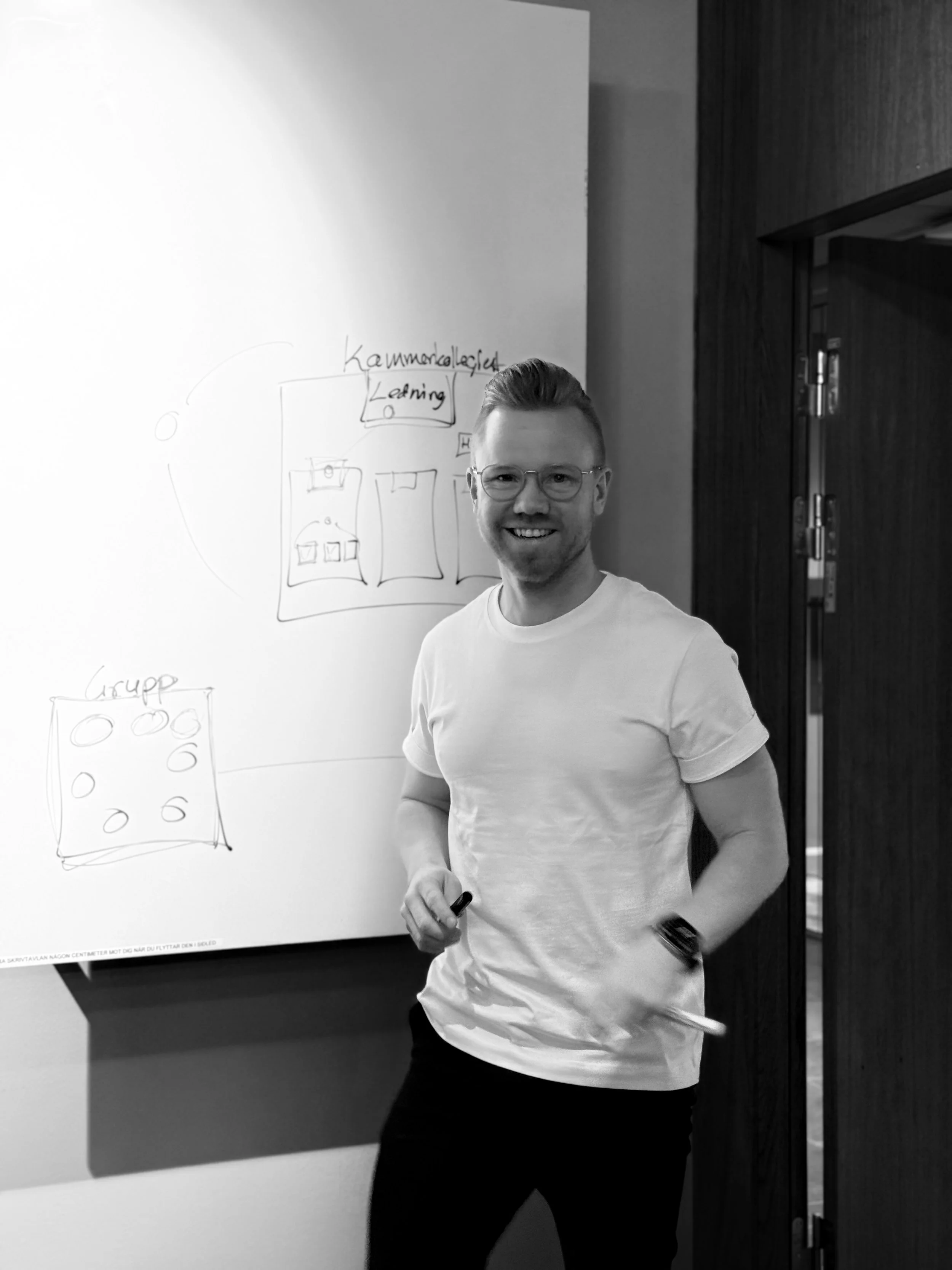 En man med glasögon och vitt T-shirt står framför en whiteboard i ett konferensrum, med teckningar och text på whiteboardet.