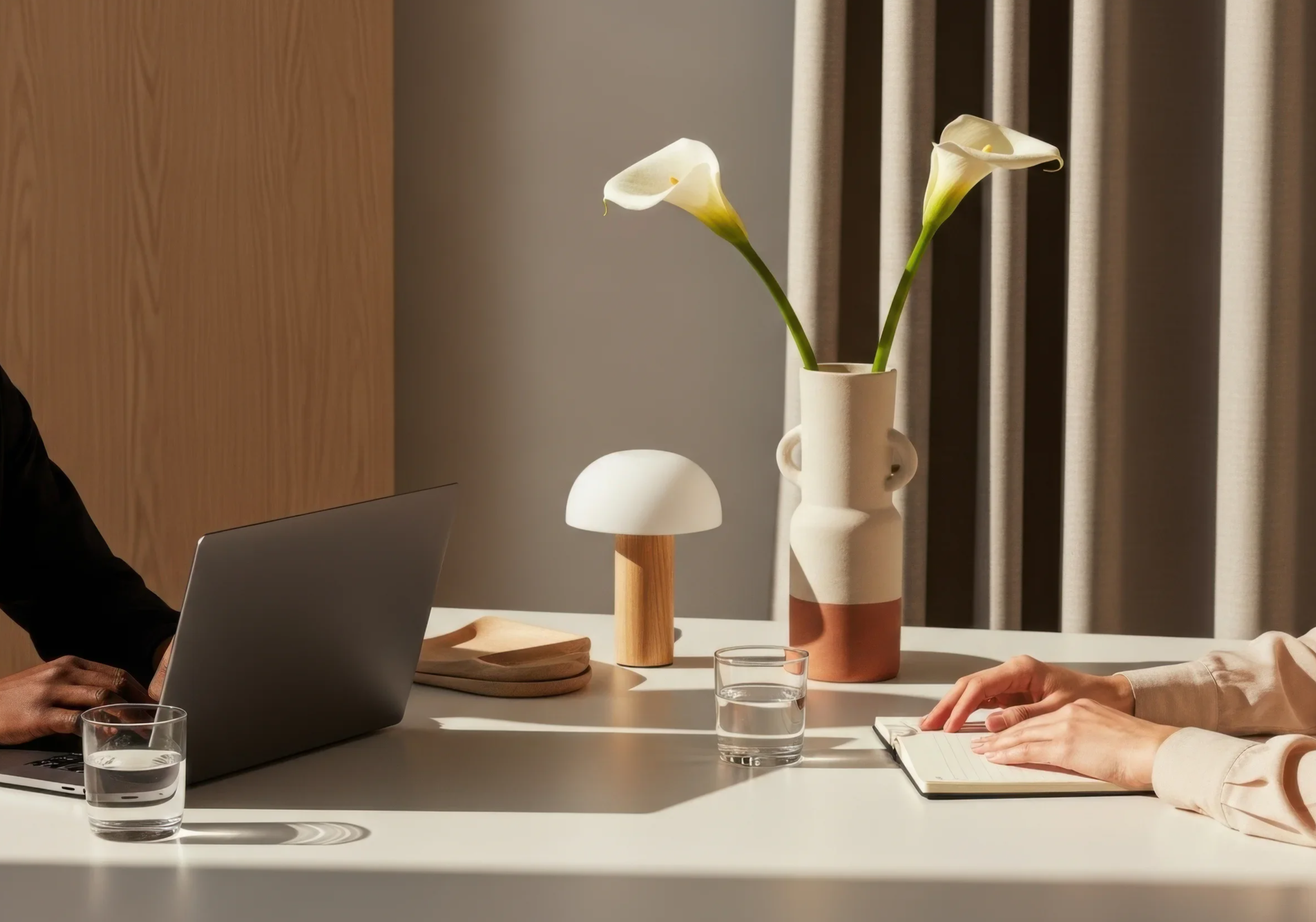 Two people sitting at a table, with one using a laptop and the other taking notes, and decorative vases with flowers on the table.